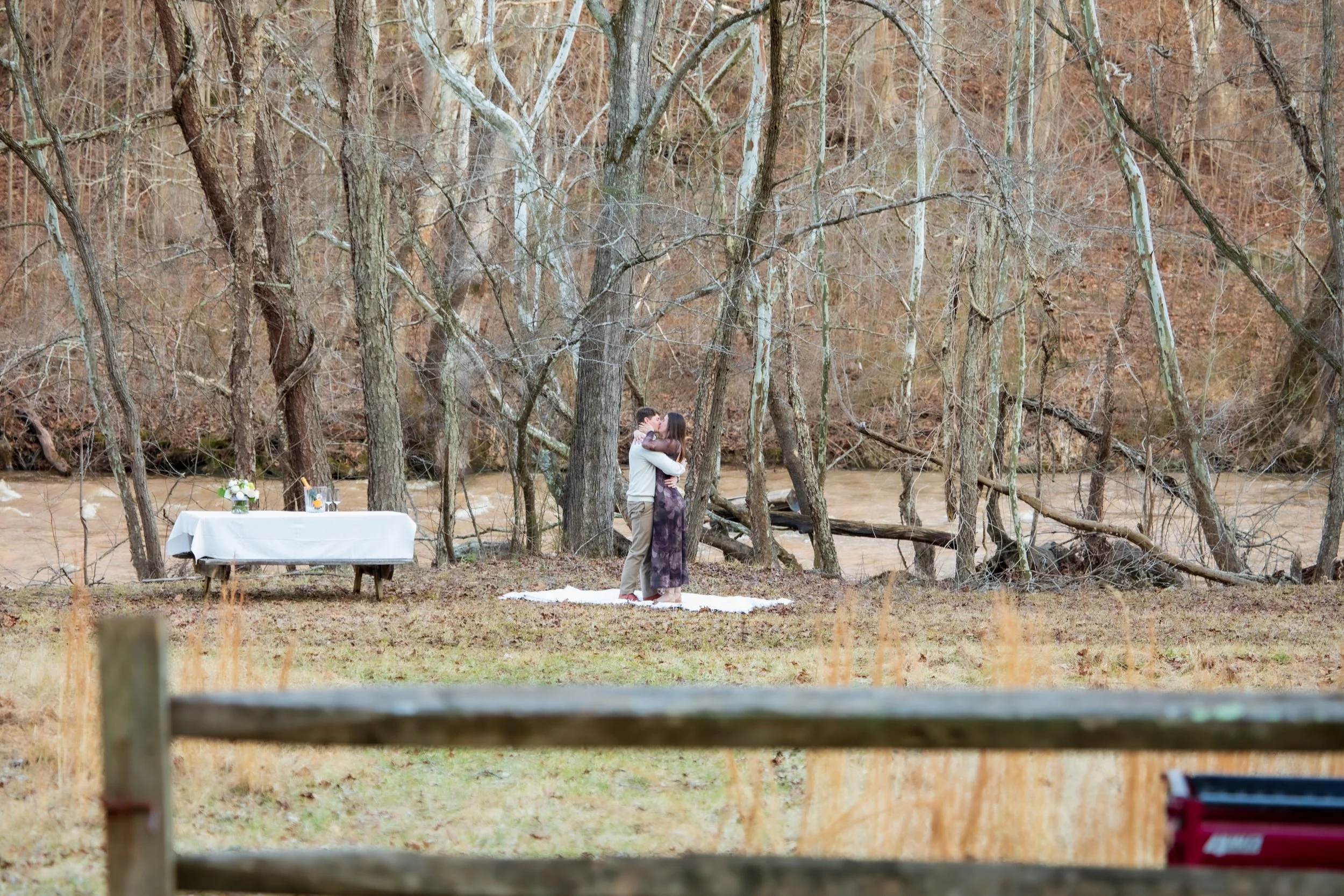 Engagement Photography by Will Locke near Richmond, VA in Montpelier. A couple hugging each other outdoors near a river, standing on a white blanket on the ground, surrounded by leafless trees in a forested area.