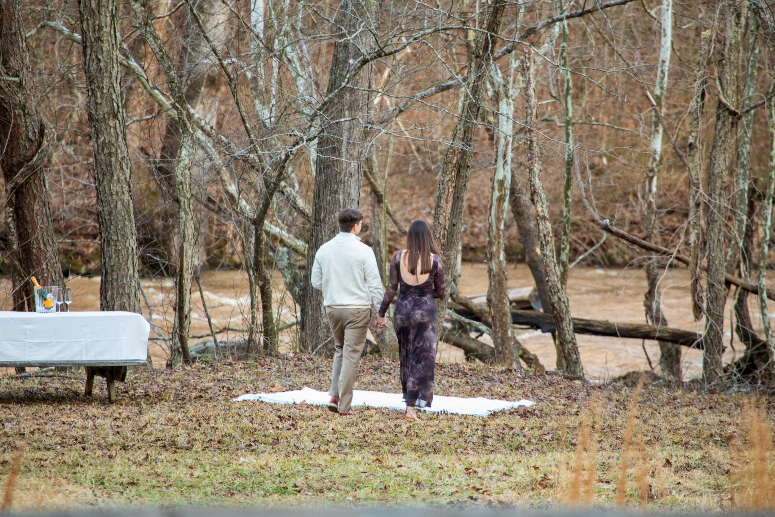 Engagement Photography by Will Locke near Richmond, VA in Montpelier. A couple walks on a white cloth on the ground near a wooded area with a stream on a fall day, with a table on the left side.