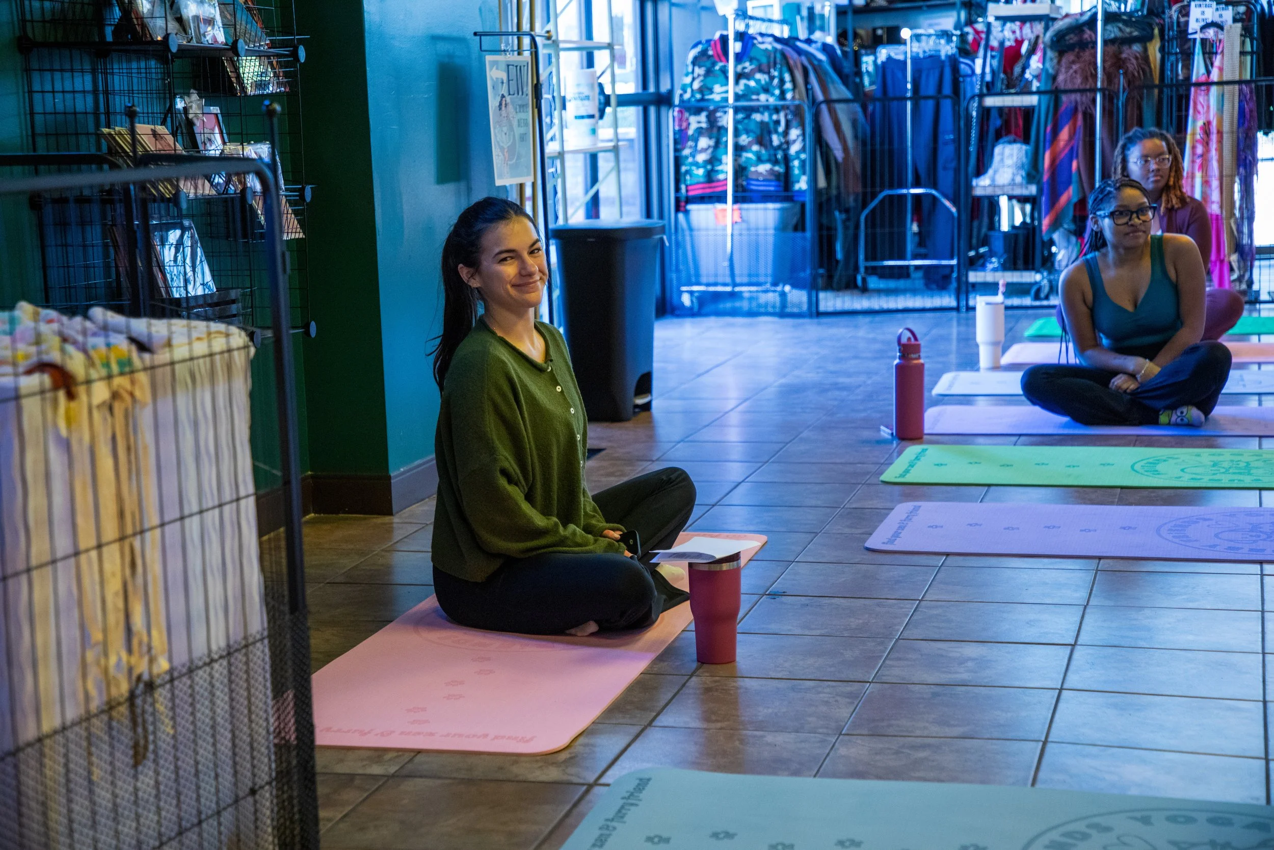 Marketing and Event Photography by Will Locke. Three women sitting cross-legged on yoga mats inside a cafe or retail store. One woman is wearing a green sweater and smiling, while the others look forward. Yoga mats and water bottles are visible.