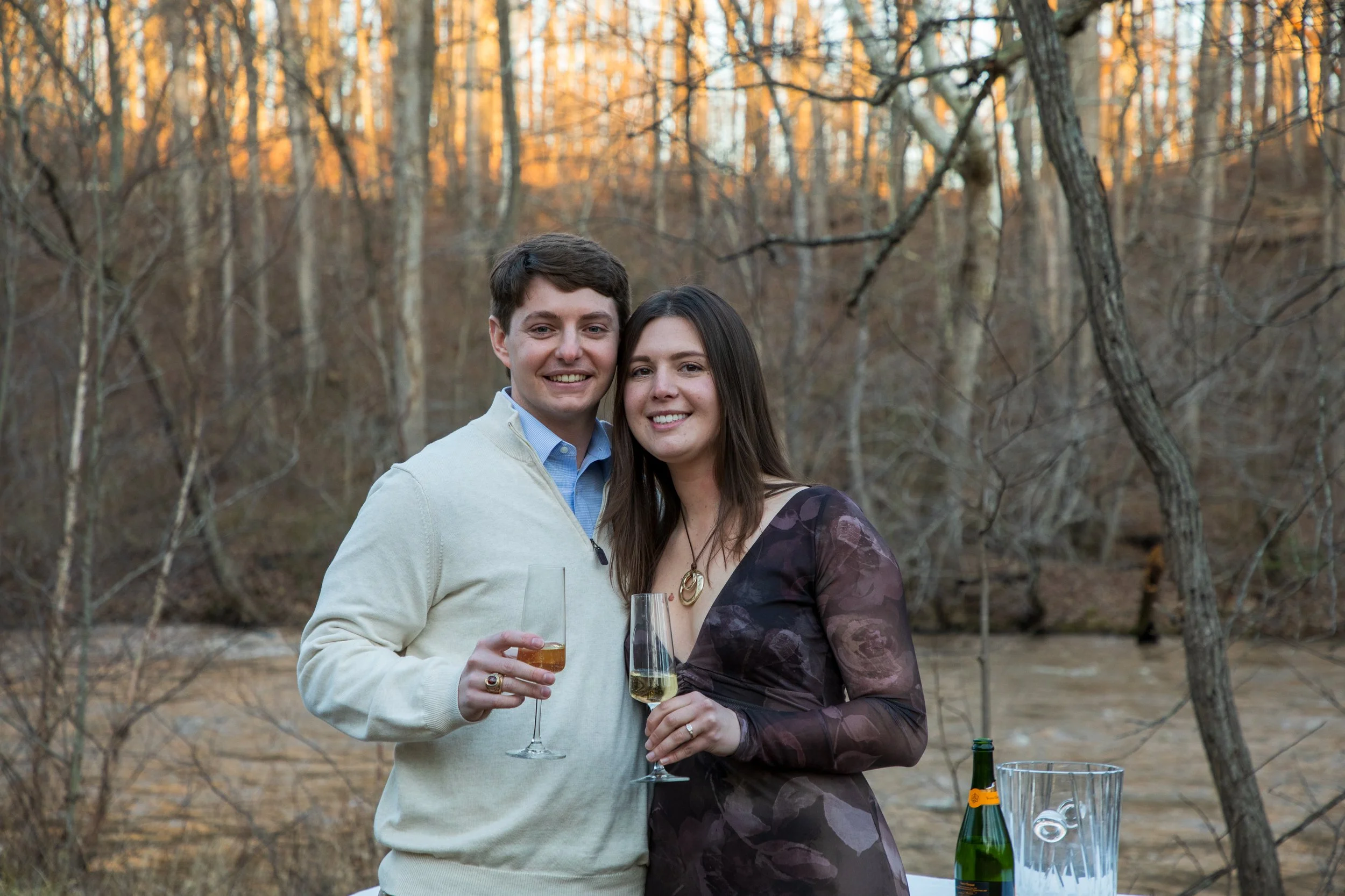Engagement Photography by Will Locke near Richmond, VA in Montpelier. A man and woman smiling and hugging while holding champagne glasses outdoors during sunset, with a table of flowers nearby.