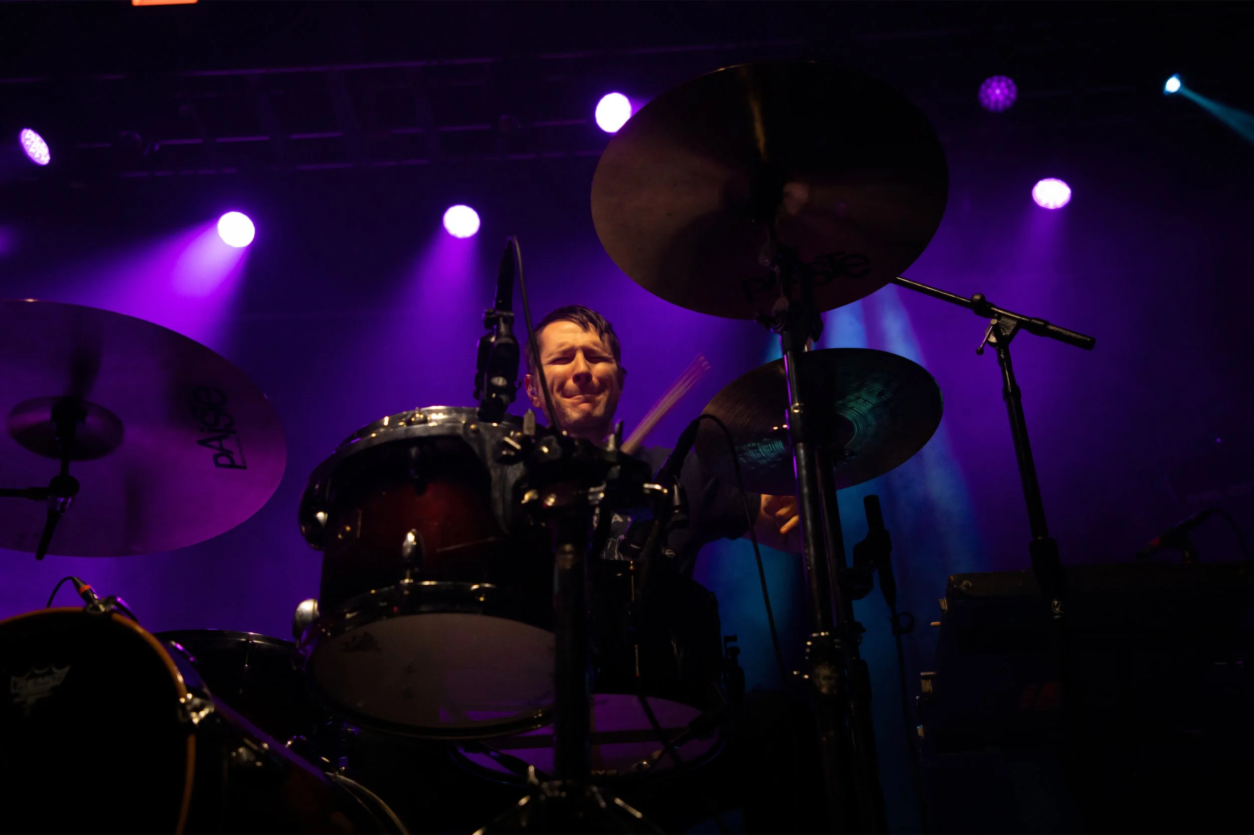 Concert Photography by Will Locke. A drummer performing on stage with purple and blue stage lights shining down.