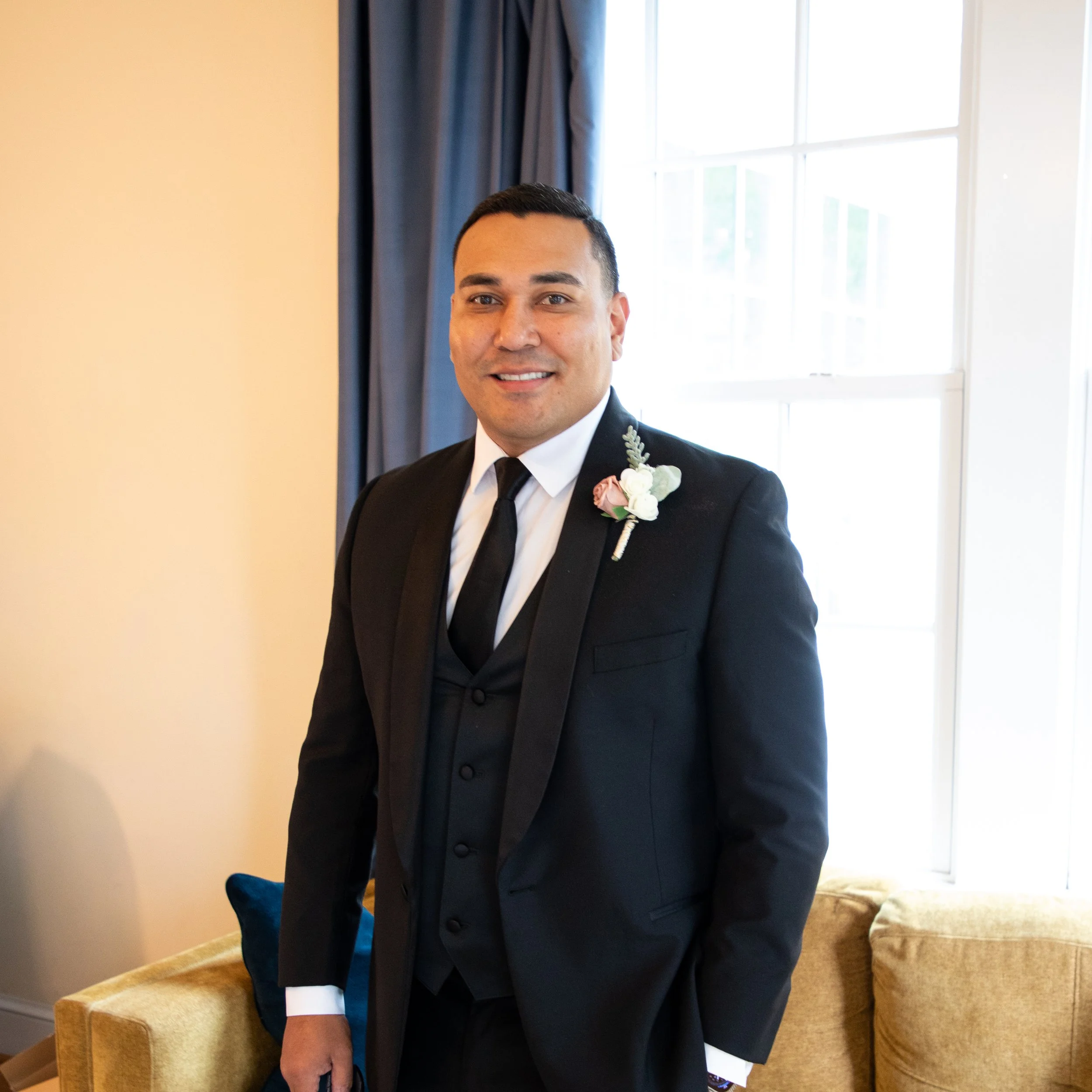 Headshot Photography by Will Locke. A man in a black suit with a white shirt, black tie, and boutonniere standing indoors near a window with blue curtains, smiling at the camera.