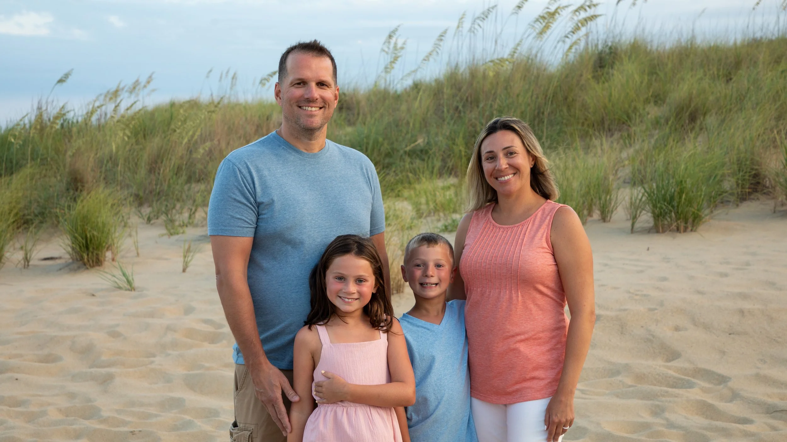 Photo & Video by Will Locke Family Photography in Virginia Beach, VA. A happy family of four standing on a sandy beach with grassy dunes behind them, smiling at the camera.