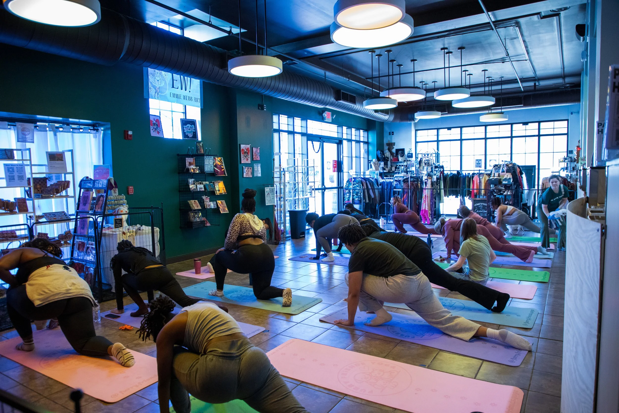 Marketing and Event Photography by Will Locke. People participating in a group yoga class inside a brightly lit retail store with large windows, clothing racks, and artwork on the walls.