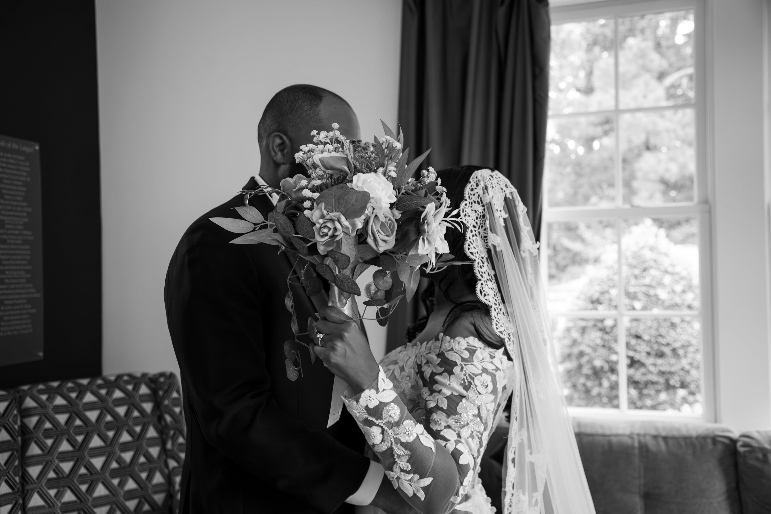 Wedding Photography by Will Locke. Black-and-white photo of a bride and groom sharing a kiss, with the bride holding a bouquet covering their faces, inside a room with large windows and curtains.