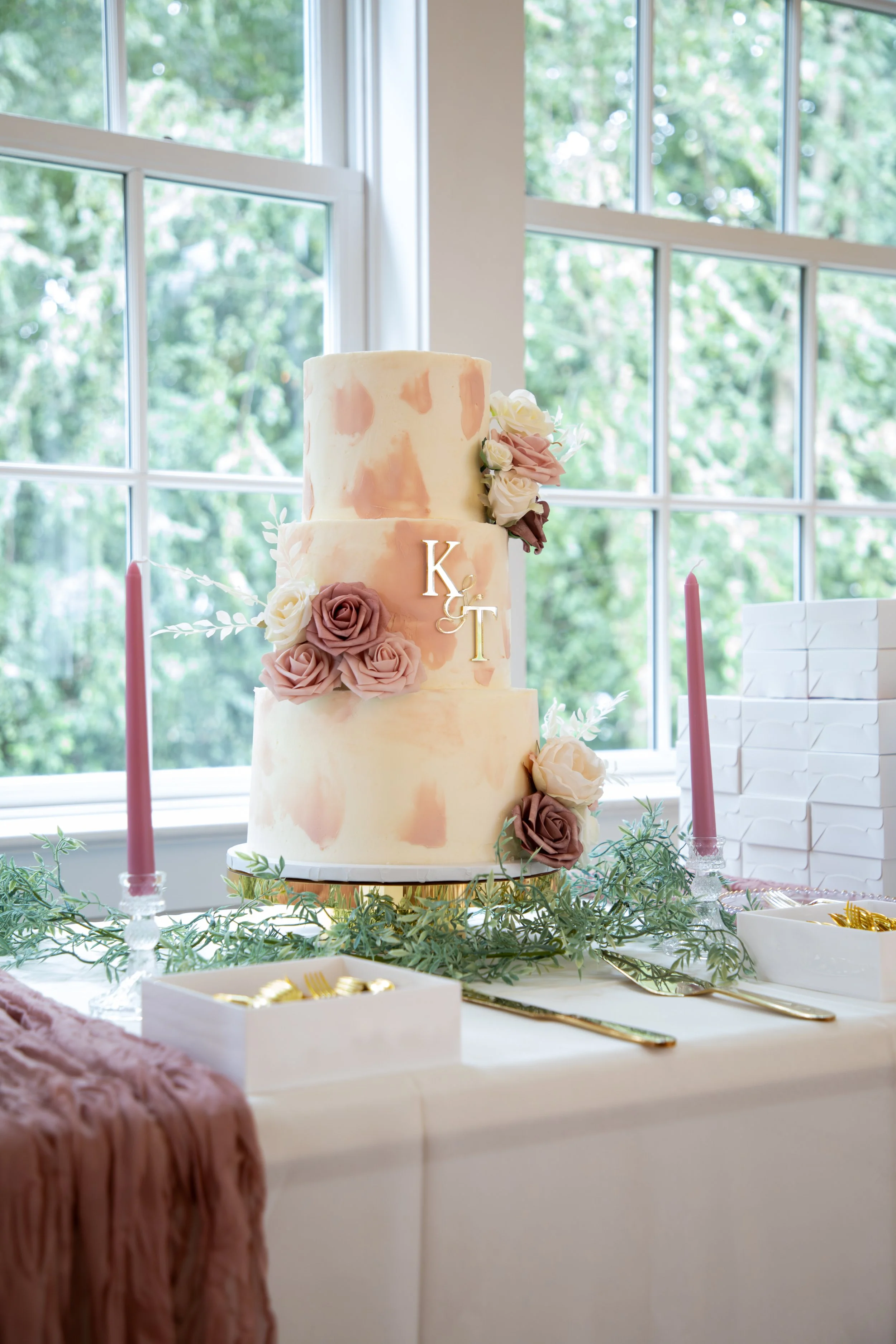 Wedding Photography by Will Locke. A three-tiered wedding cake with pink, beige, and white marbled frosting, decorated with pink and white roses and greenery, placed on a table with pink candles, gold utensils, and a pink cloth.