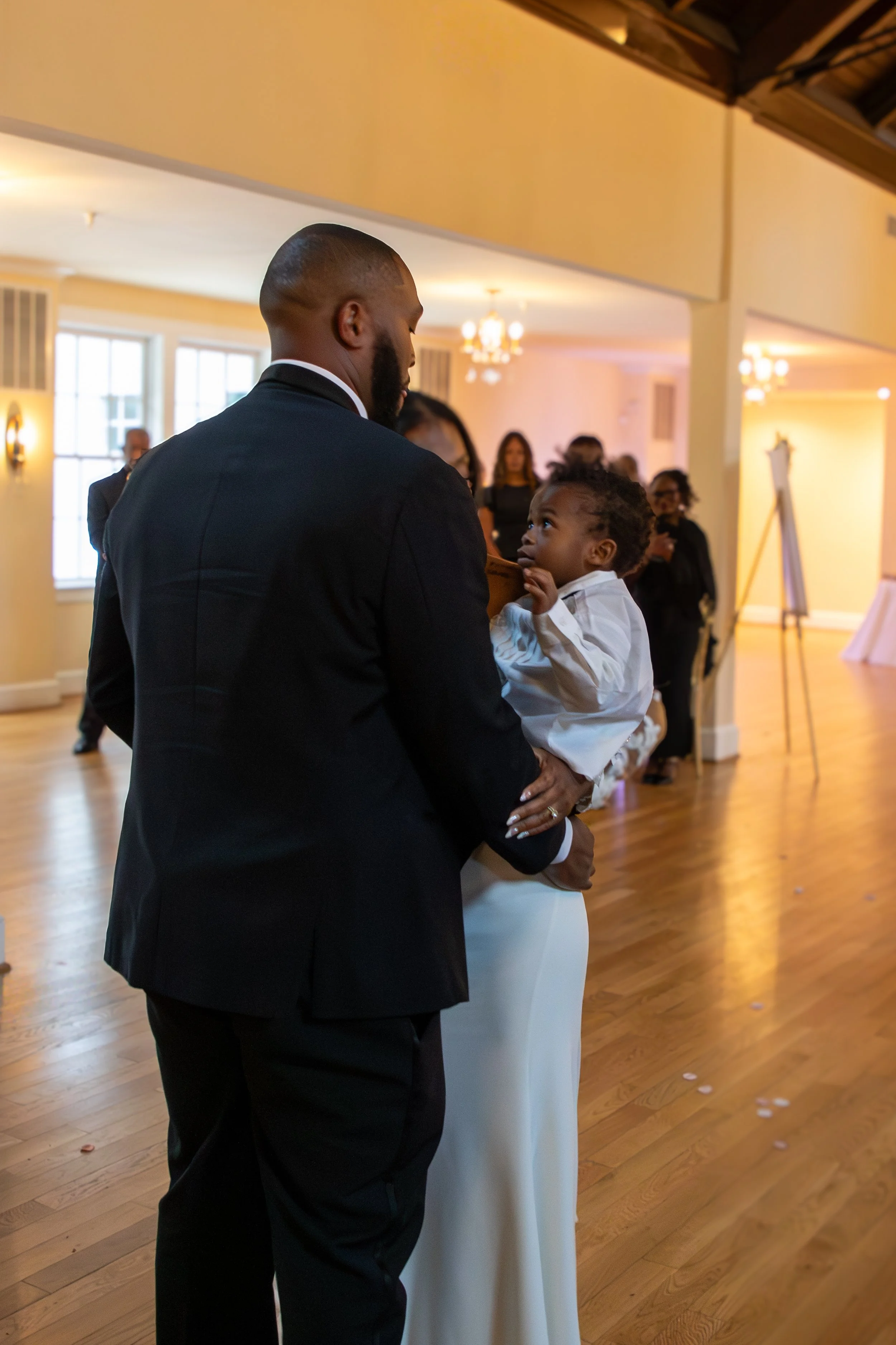 Photo & Video by Will Locke Wedding Photography at the Woman's Club of Portsmouth in Virginia. A man holding a young girl in a wedding dress during a wedding reception in a decorated indoor venue.