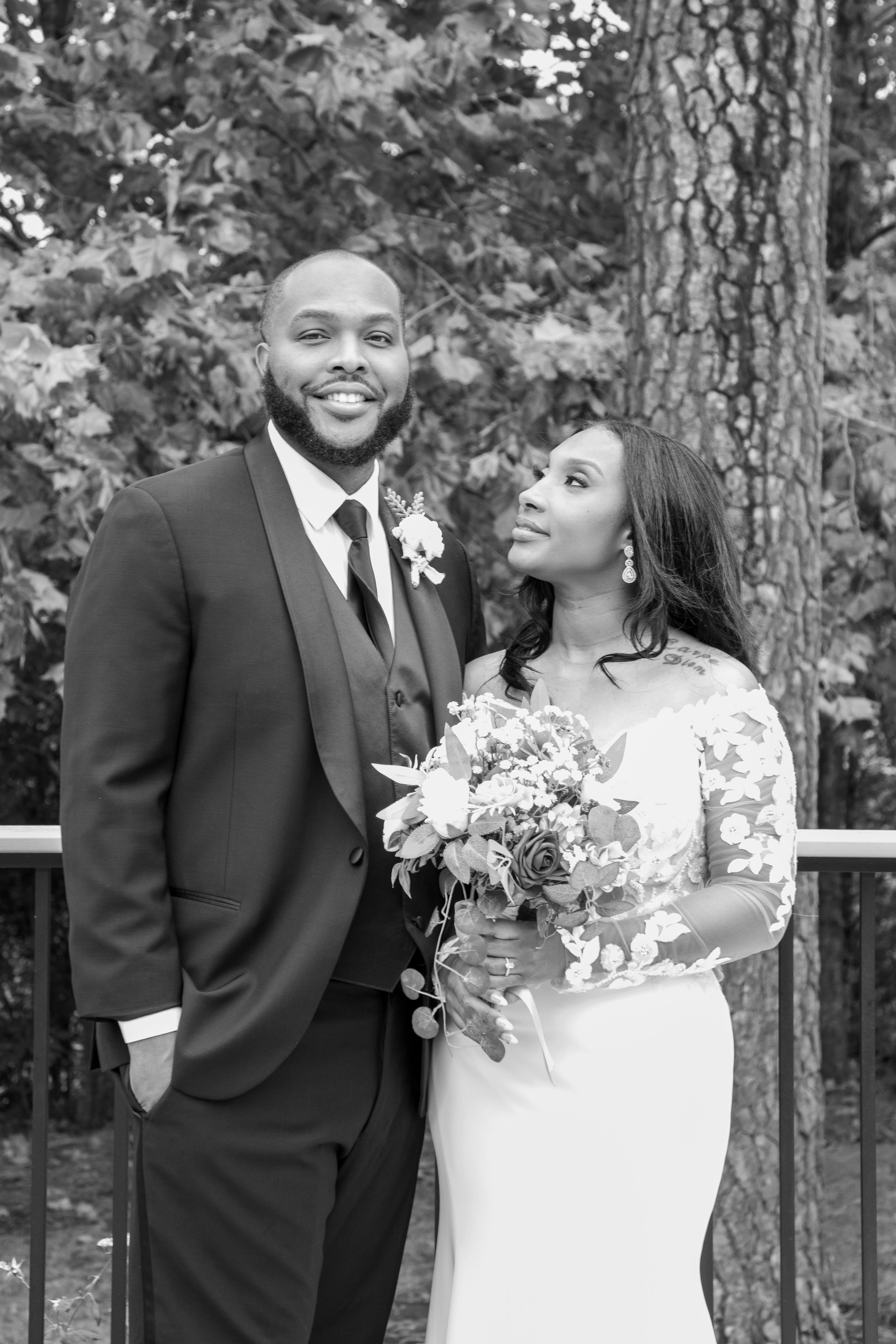 Photo & Video by Will Locke Wedding Photography at the Woman's Club of Portsmouth in Virginia. A black and white photo of a newlywed couple outdoors, standing in front of trees, with the bride holding a bouquet and looking at the groom.