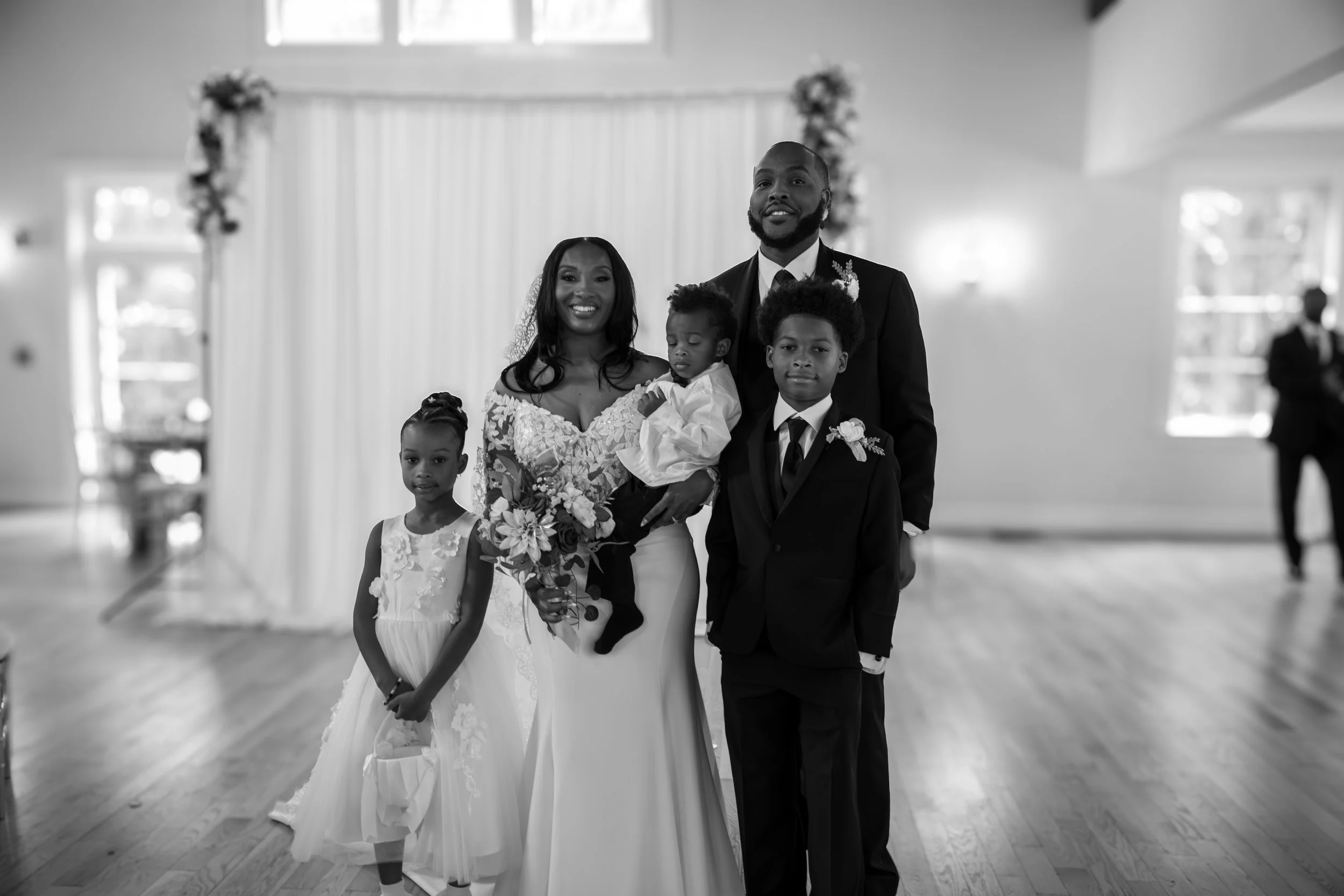 Photo & Video by Will Locke Wedding Photography at the Woman's Club of Portsmouth in Virginia. A family dressed in formal wedding attire stands together inside a decorated hall for a wedding ceremony. 