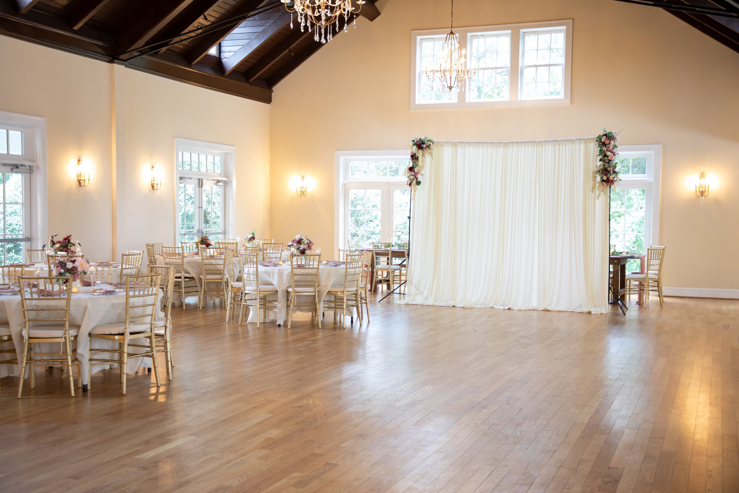 Wedding Photography by Will Locke. A decorated wedding reception area inside a bright hall with large windows, gold chairs, white tablecloths, and floral centerpieces, with a white curtain backdrop and crystal chandeliers.