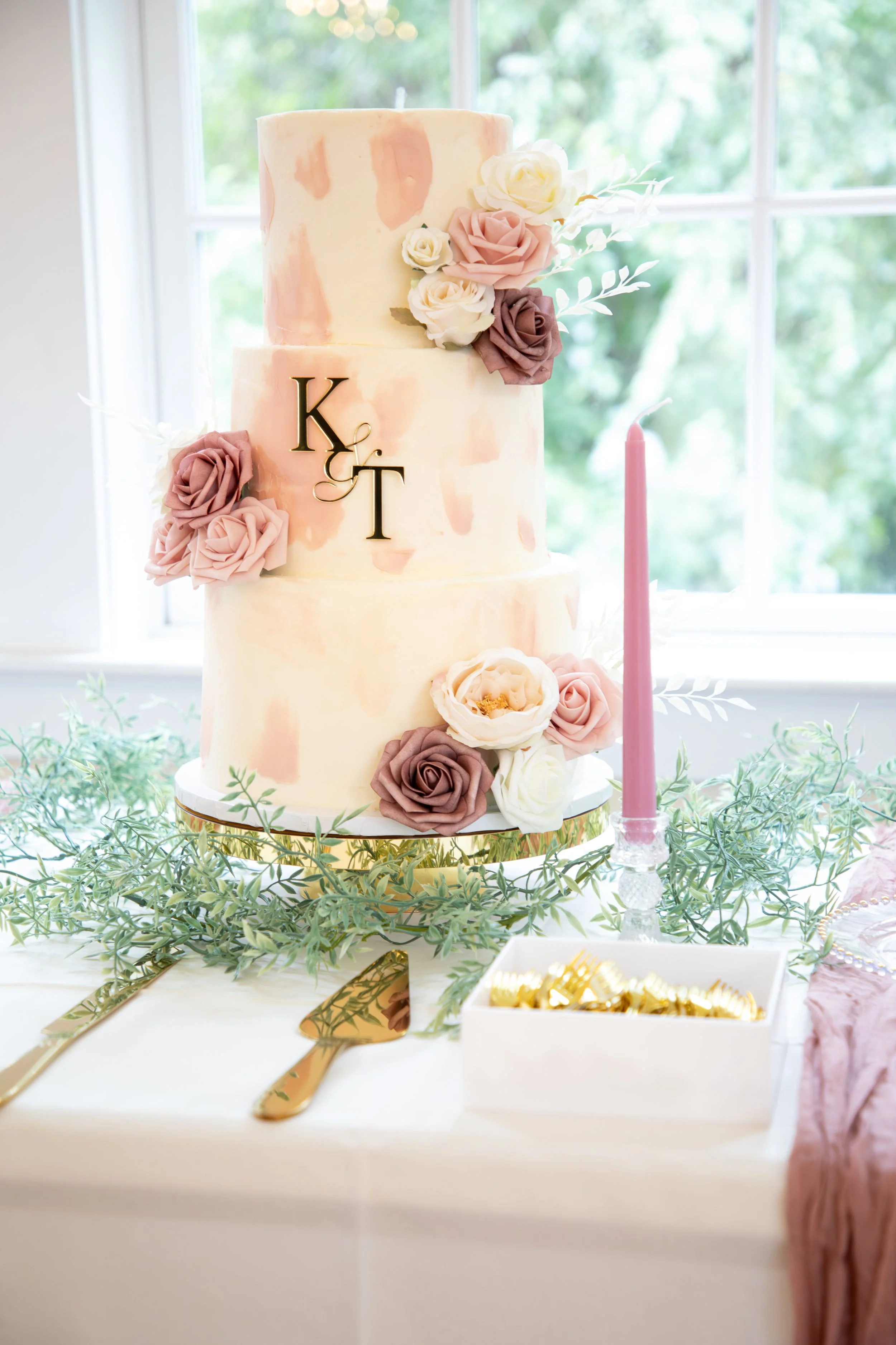 Wedding Photography by Will LThree-tier wedding cake decorated with pink, white, and mauve flowers, with gold monogram initials 'K & T' on the middle tier, set on a gold cake stand surrounded by greenery and a pink taper candle, in front of a window.