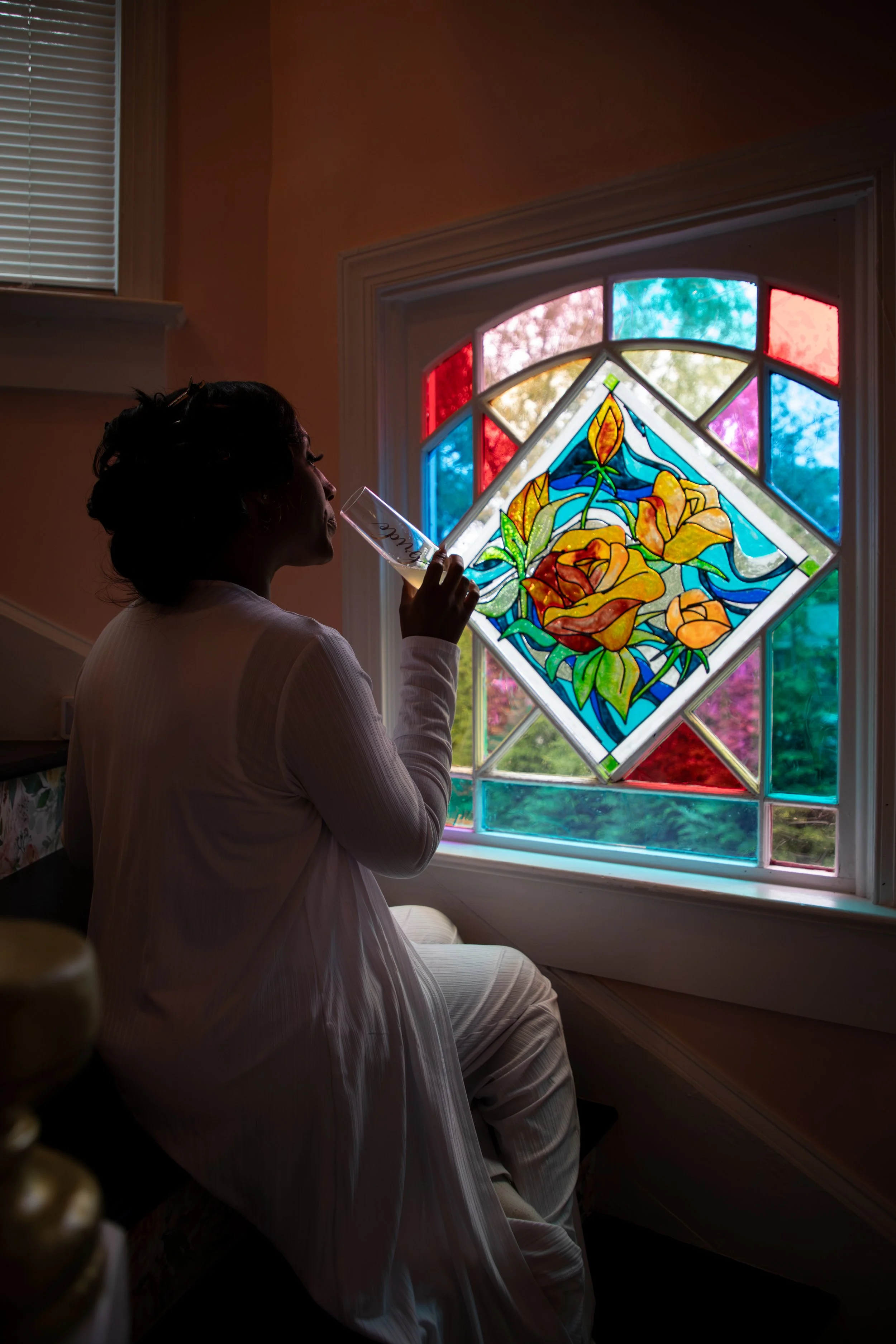 Photo & Video by Will Locke Bridesmaid Wedding Photography at The Bridgerton House in Portsmouth, VA. Woman sitting by a stained glass window with floral and geometric designs, drinking from a glass.