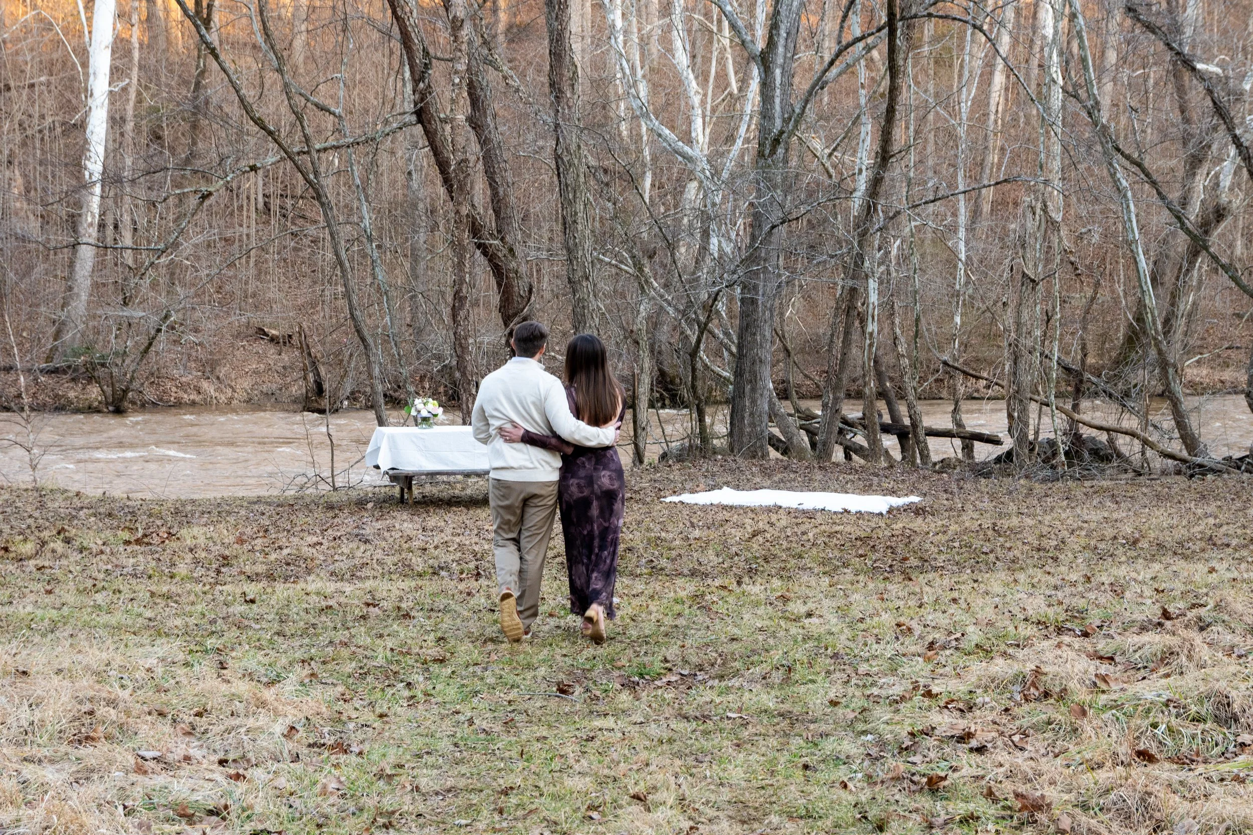 Engagement Photography by Will Locke near Richmond, VA in Montpelier. A couple walks to a white cloth on the ground near a wooded area with a stream on a fall day, with a table on the left side with white tablecloth.