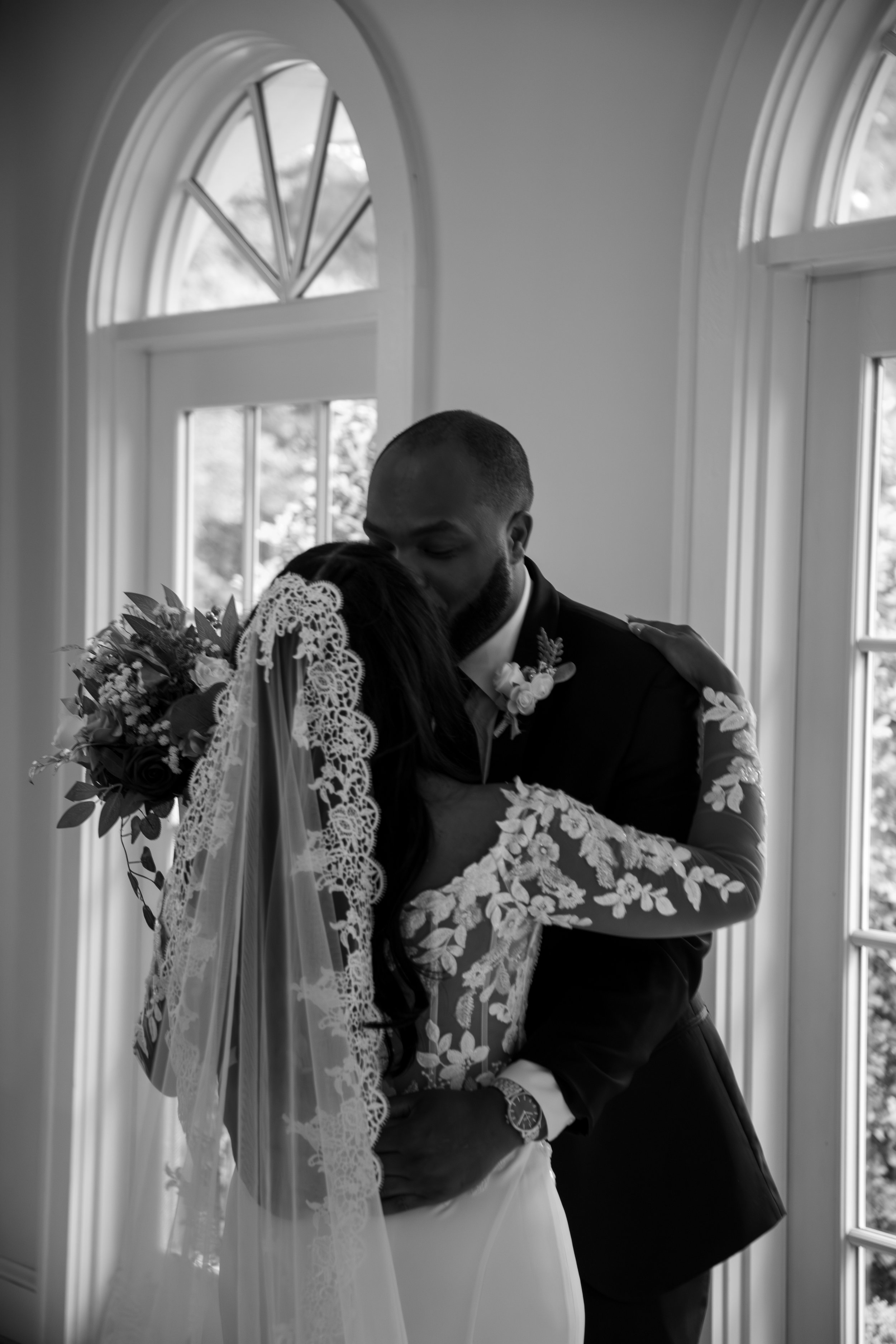 Wedding Photography by Will Locke. A black and white photo of a newlywed couple embracing indoors near windows, with the bride holding a bouquet and wearing a lace wedding dress and veil, and the groom in a suit.