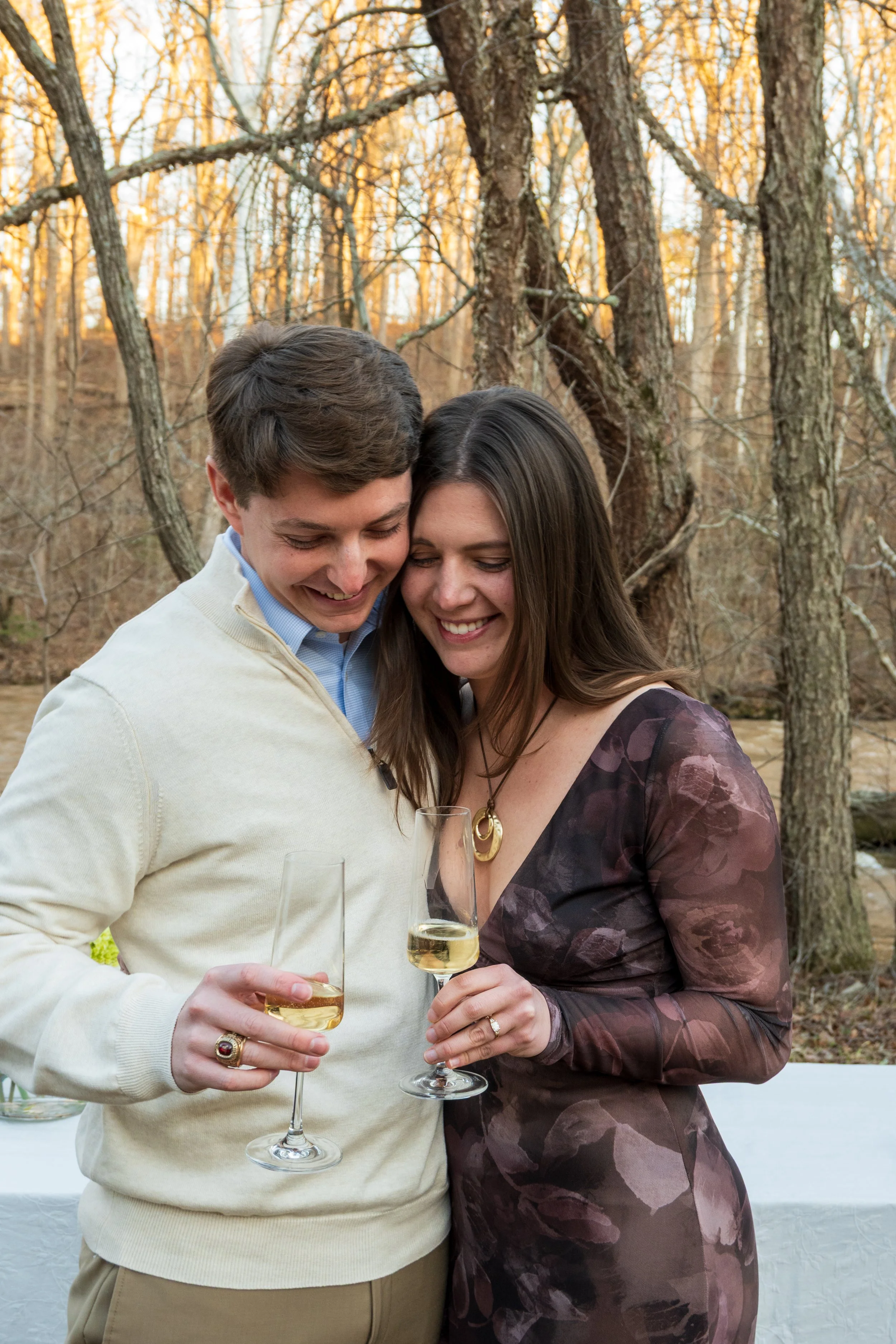 Engagement Photography by Will Locke near Richmond, VA in Montpelier. A man and woman smiling and hugging while holding champagne glasses outdoors during sunset, with a table of flowers nearby.
