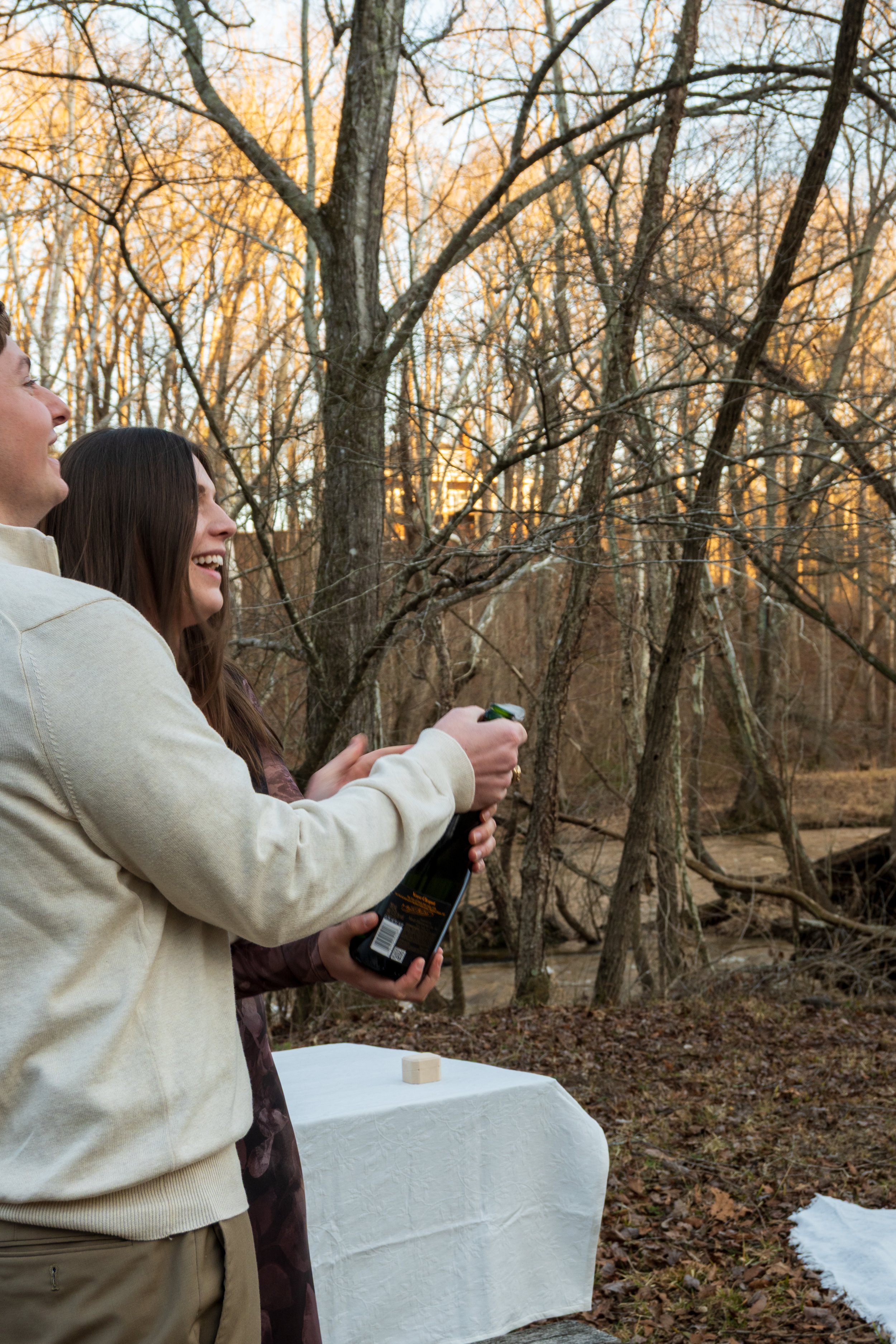 Engagement Photography by Will Locke near Richmond, VA in Montpelier. A man and woman pop a champagne bottle outdoors during sunset, with a table with white tablecloth nearby.