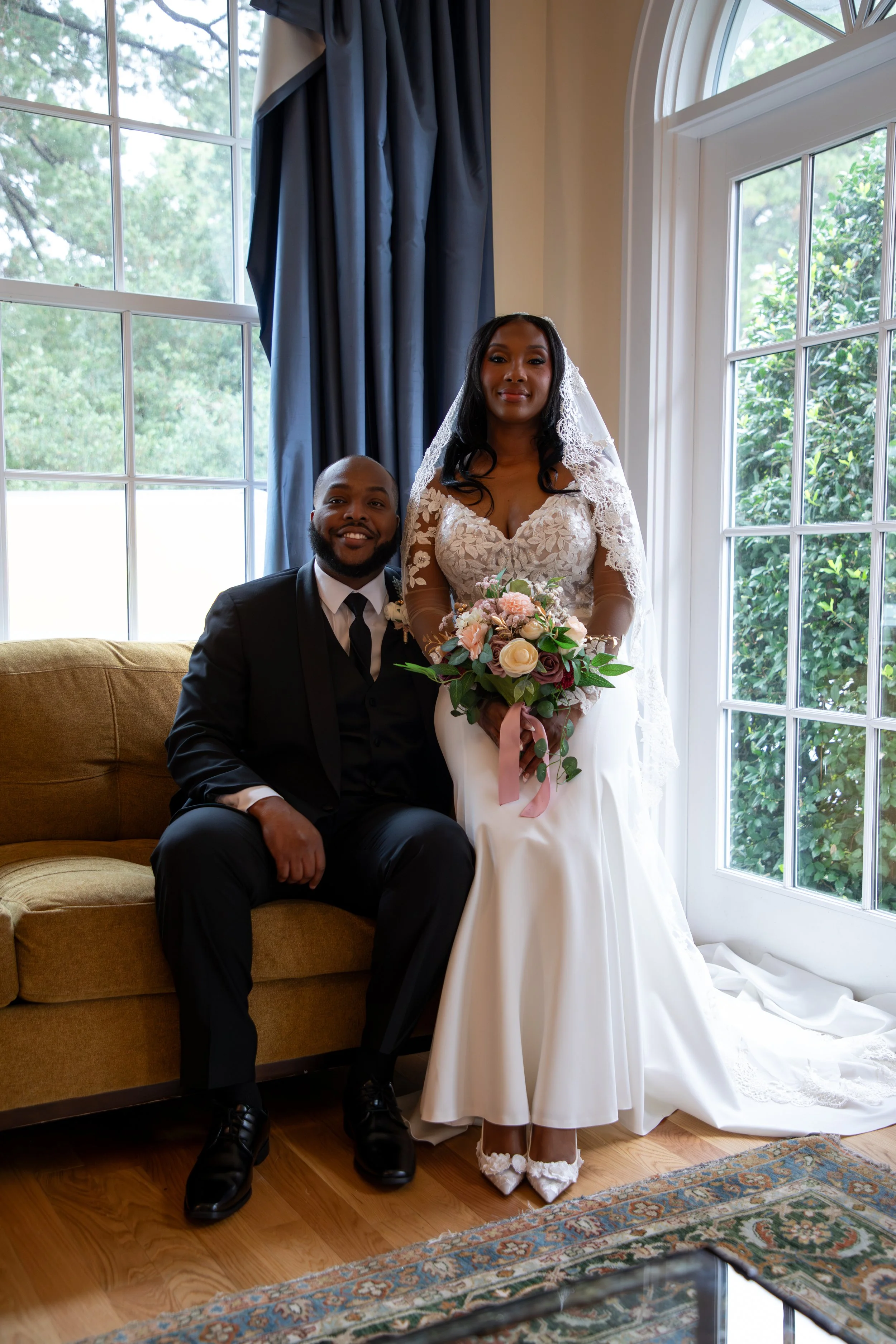 Photo & Video by Will Locke Wedding Photography at the Woman's Club of Portsmouth in VA. A bride in a white wedding dress holding a bouquet and a groom in a black suit sitting beside her on a brown sofa in front of large windows with blue curtains.