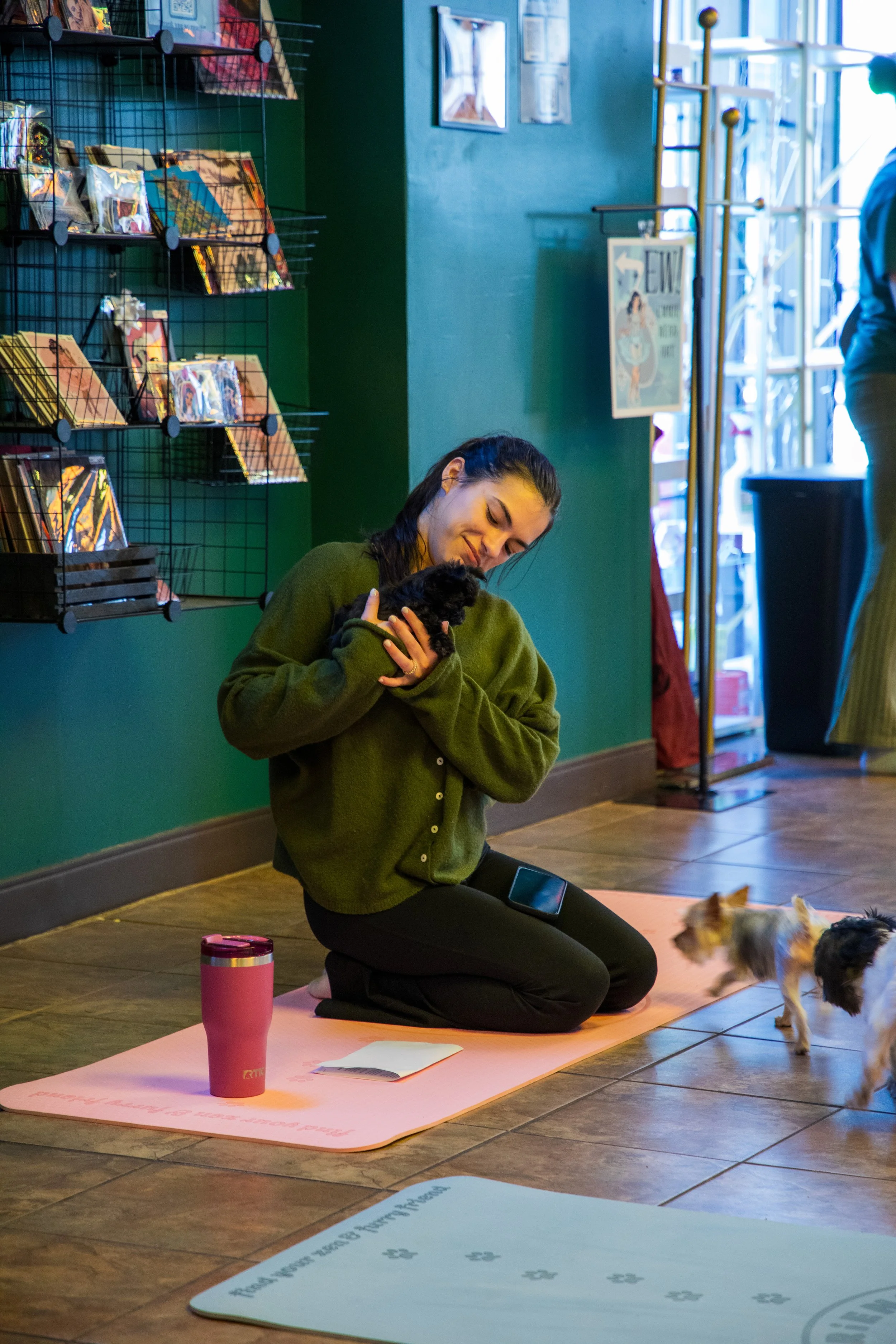 A woman kneeling on a pink yoga mat inside a pet store, holding a small black puppy, with two other small dogs approaching. Marketing and Event Photography by Will Locke. 