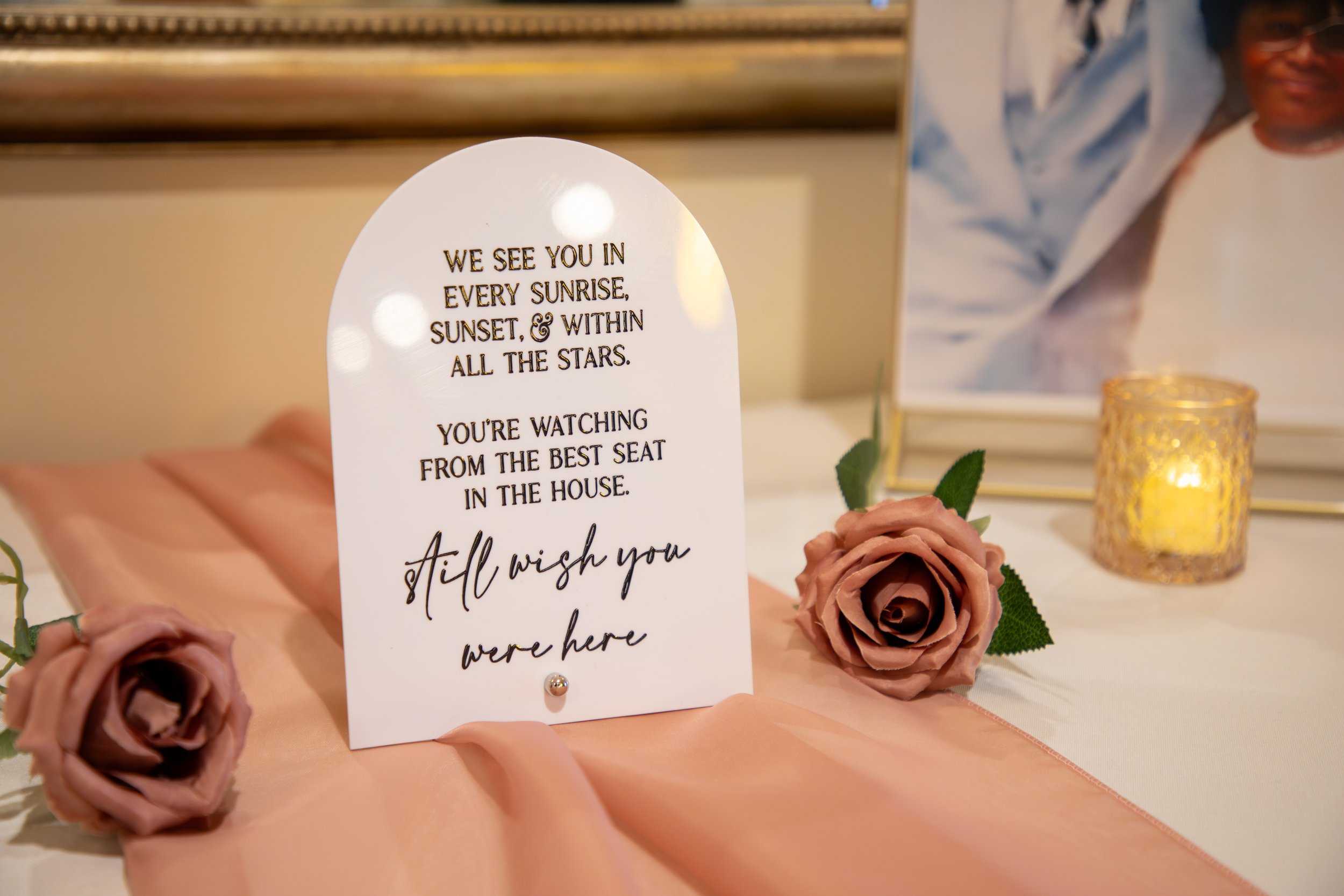 Wedding Photography by Will Locke. A decorative table display featuring a white card with a heartfelt message, pink roses, a photo frame with a person, and a lit candle in a gold holder.