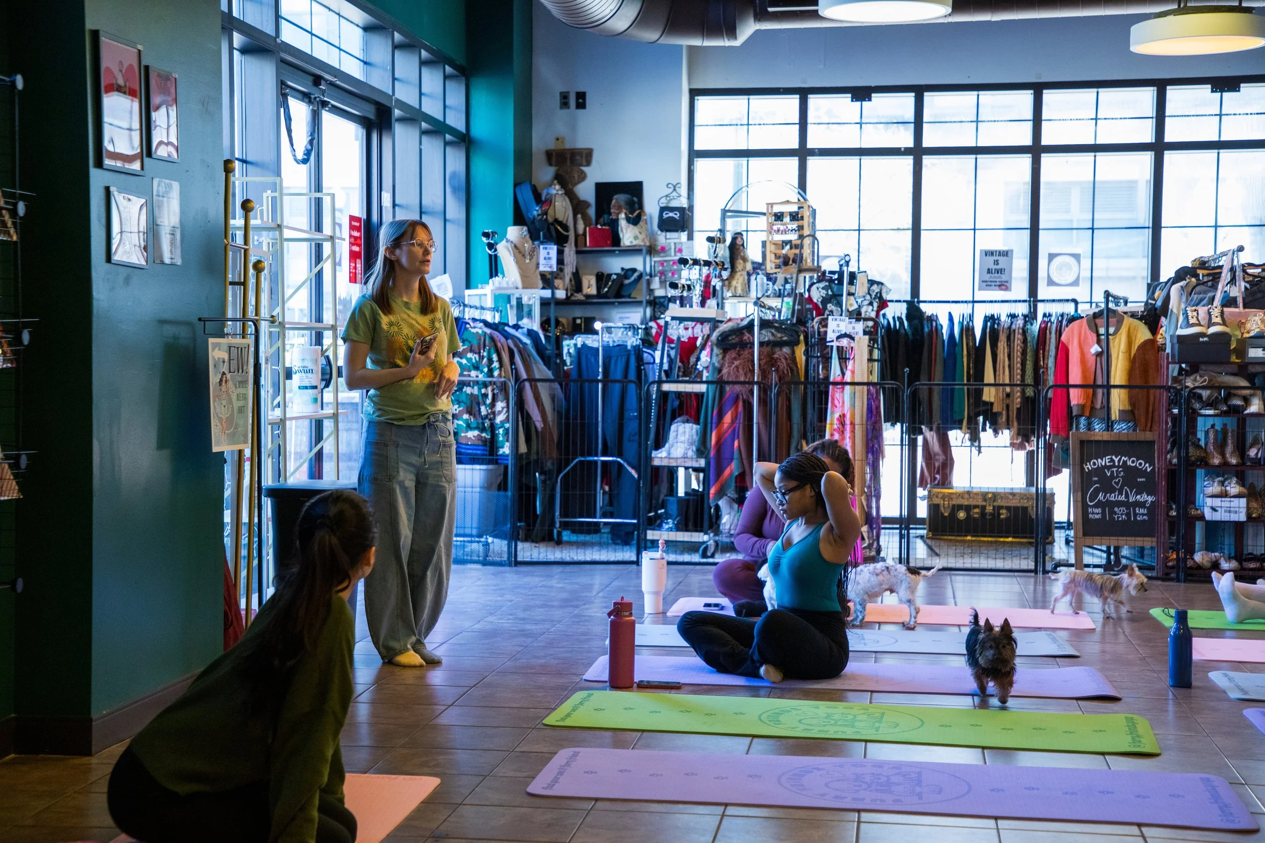 Marketing and Event Photography by Will Locke. Group of people participating in a yoga or meditation class inside a store with clothing and accessories, some sitting on yoga mats and a woman standing near a clothing rack.