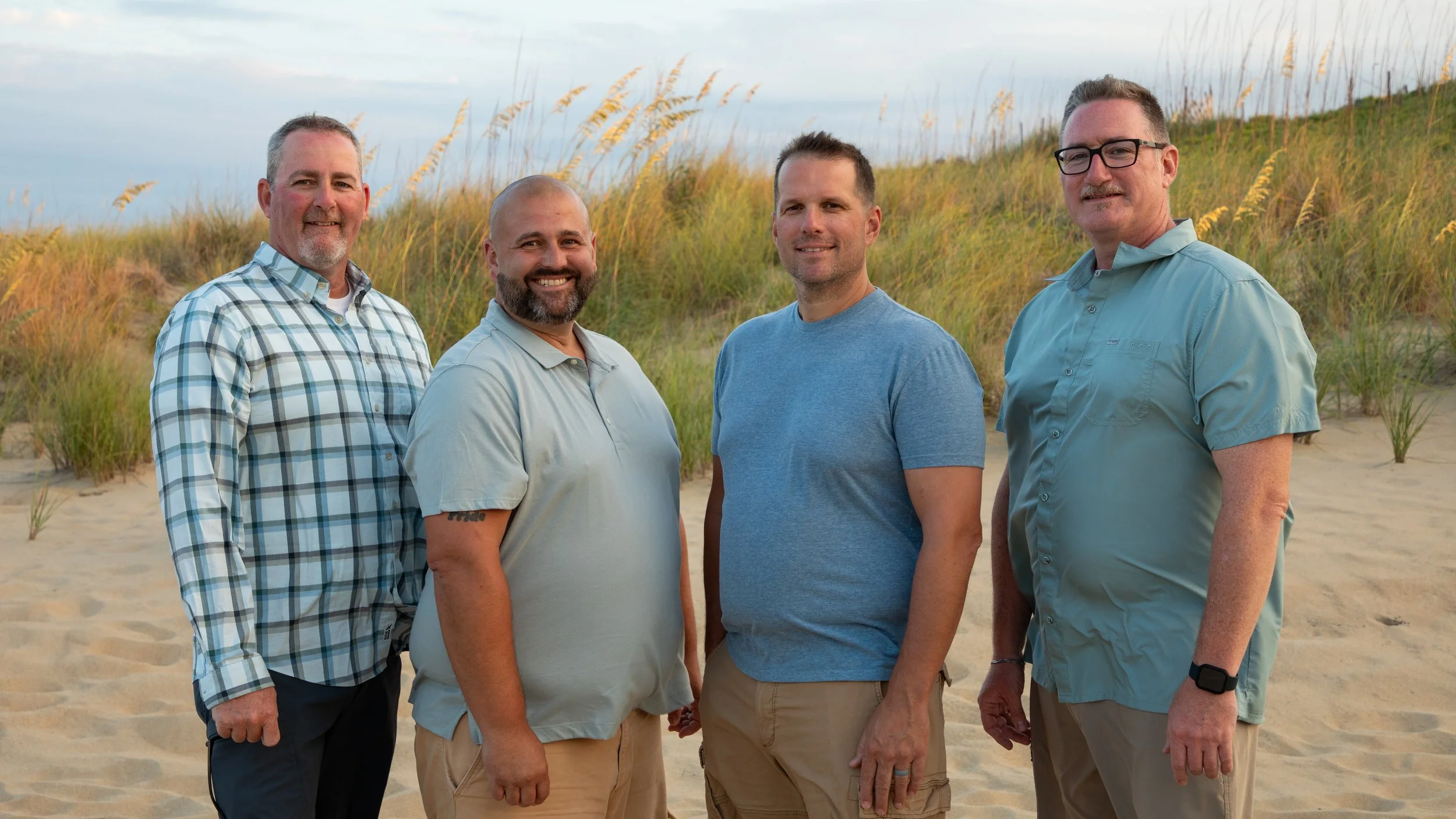 Photo & Video by Will Locke Family Photography in Virginia Beach, VA. Four men standing on a sandy beach with grass dunes in the background, smiling at the camera.