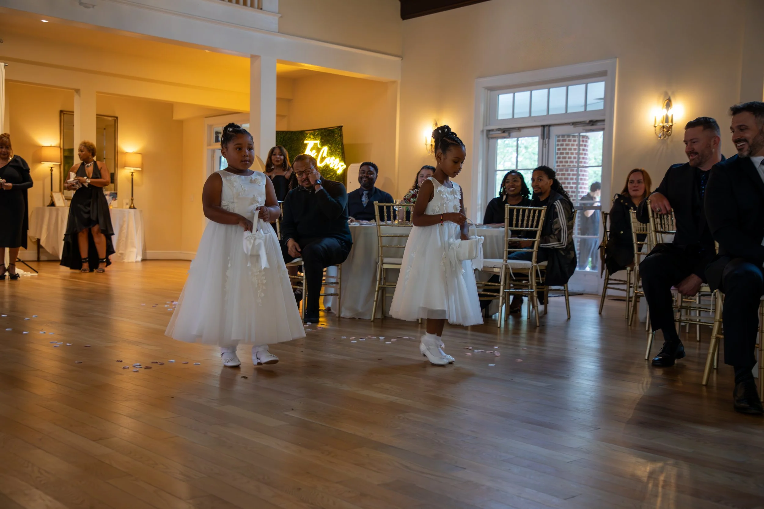 Wedding Photography by Will Locke. Two young girls in white dresses and white shoes walk across a wooden floor at a wedding reception, surrounded by seated guests in formal attire, with some standing and chatting in the background.