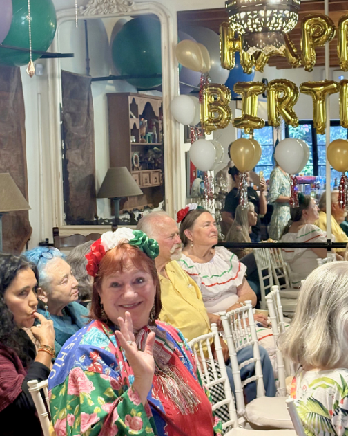 A group of women at a birthday party, decorated with balloons and gold balloon letters spelling "Happy Birthday", with several women seated and enjoying the celebration.