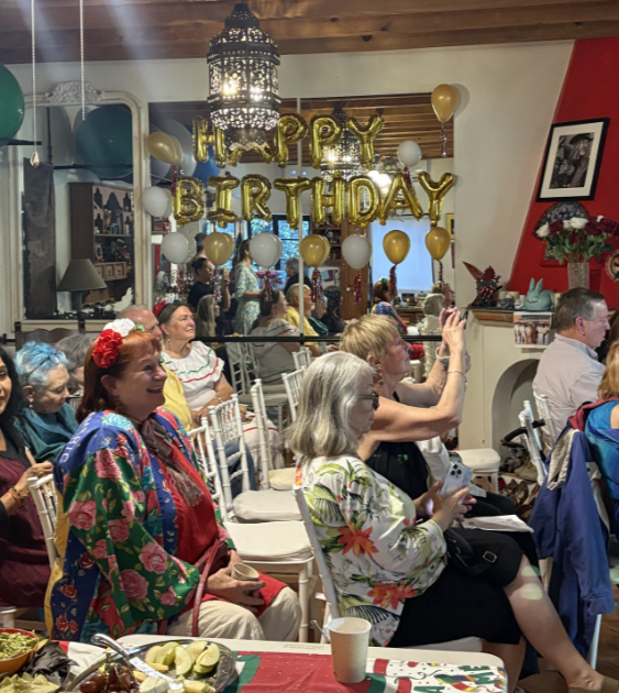 People celebrating a birthday party with balloons and a 'Happy Birthday' banner, gathered indoors with decorations and food.