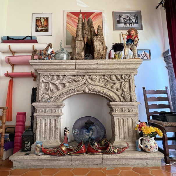 Decorative fireplace with various ornaments and artwork on mantel, including a large stone sculpture of Sagrada Familia, dolls, a pot, framed pictures, a flower vase, and garments on for sale.