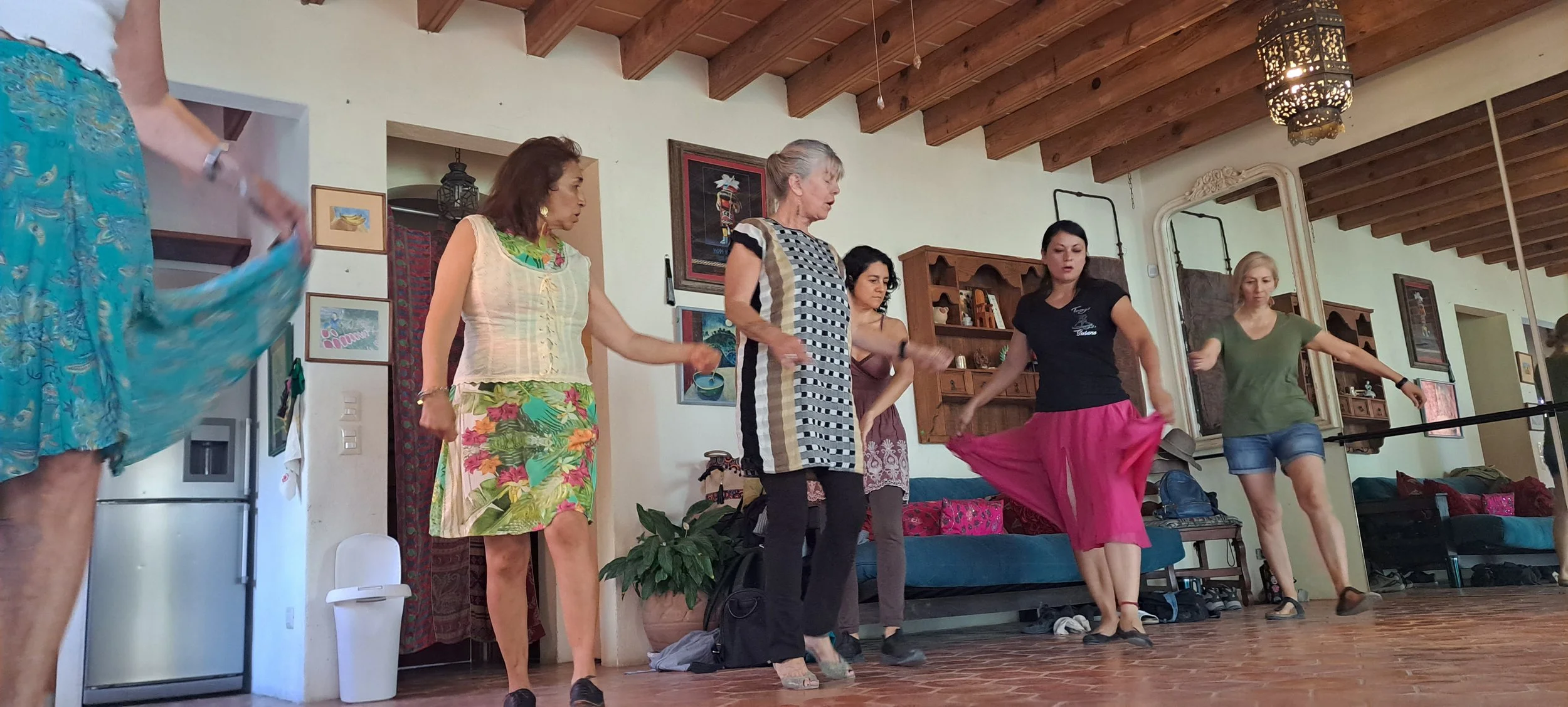 Group of women standing in a line while wearing colorful skirts or dresses, possibly dancing or participating in a cultural activity, in a room with a high wooden ceiling and framed artwork on the wall.