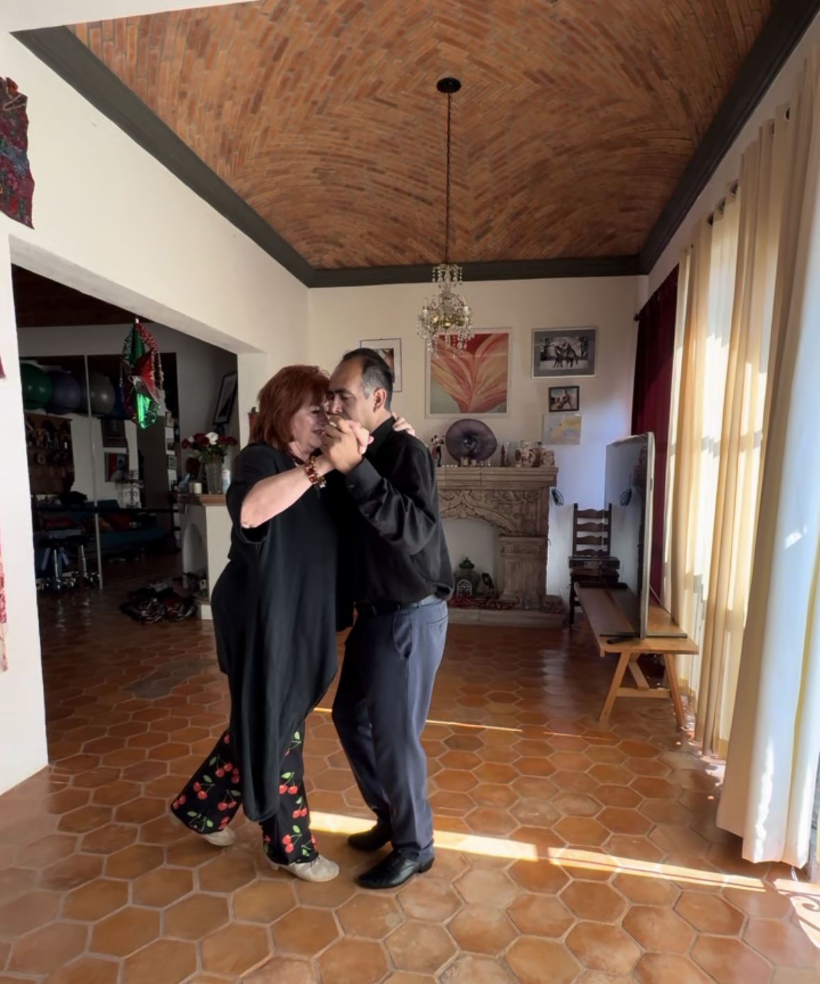 A woman and a man dancing closely in a warmly lit living room with a vaulted brick ceiling, a chandelier, curtains, and various artwork on the wall.