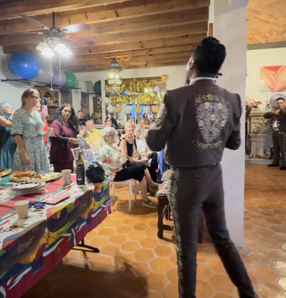 A person in a black jacket with decorative embroidery on the back is performing for a crowd at a birthday party. The audience includes elderly and middle-aged people, some sitting and some standing, smiling and enjoying the event. Decorations include a 'Happy Birthday' banner with balloons, a table with food, and colorful tablecloths. The setting is a cozy, rustic room with wooden ceilings and tiled floors.