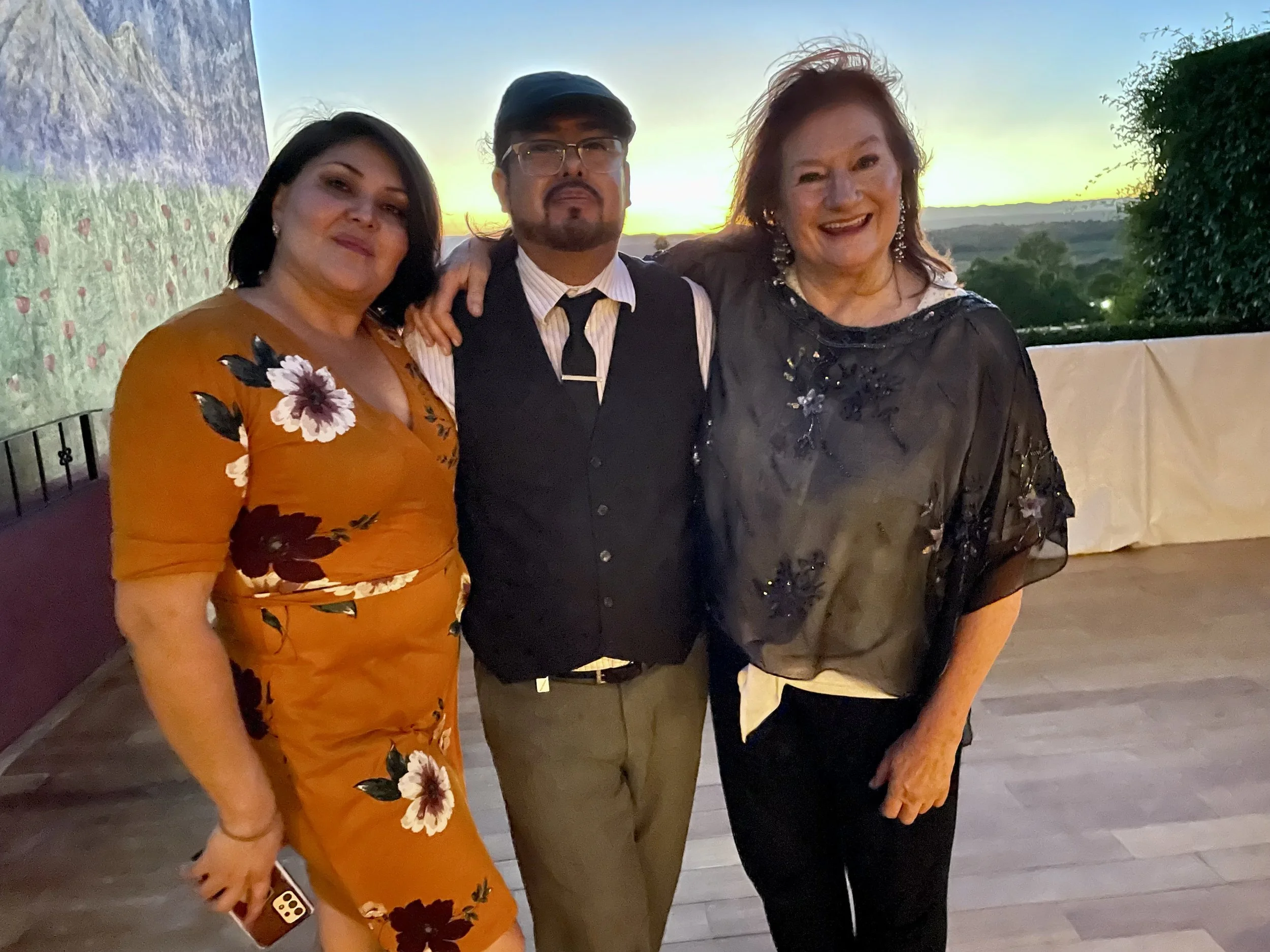 Three people standing together outdoors during sunset, smiling and dressed in formal and semi-formal clothing.