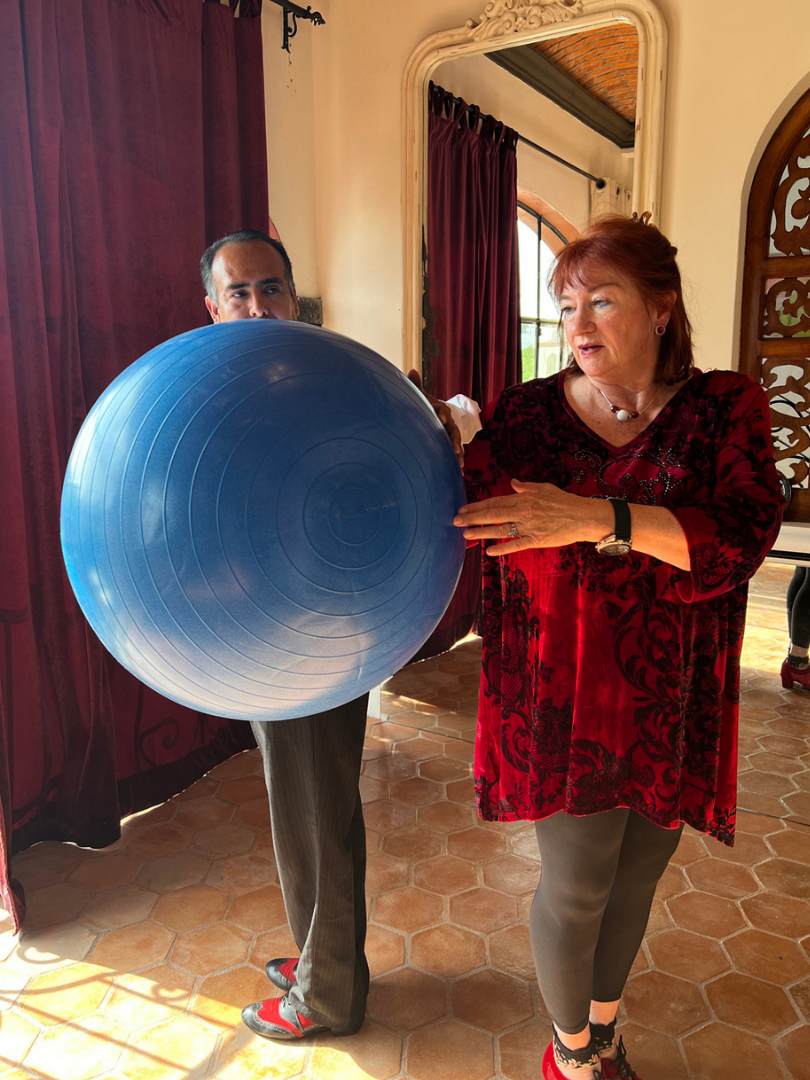 A woman with red hair holding a blue exercise ball, with a man standing partially behind it. They appear to be indoors in a room with red curtains, a mirror, and decorative wooden panels.