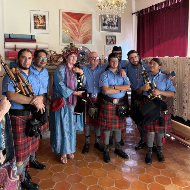 A group of seven people, six men and one woman, dressed in traditional Scottish attire with kilts, standing indoors. They are holding bagpipes and smiling for the photo in a room decorated with colorful artwork, a chandelier, and red curtains.