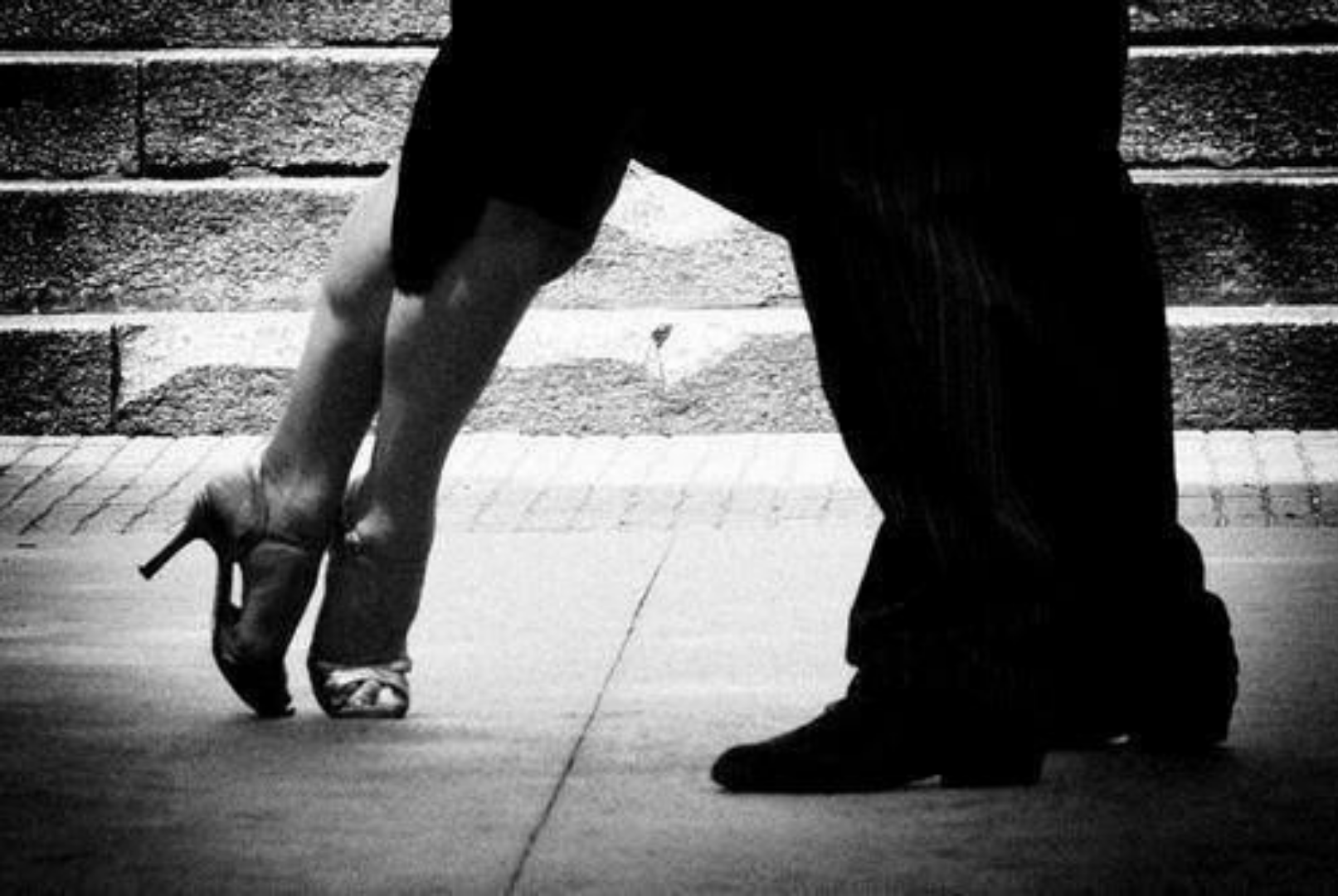 Silhouette of a person in high-heeled shoes bending down, with another person kneeling in front of them on a sidewalk, against a brick wall background.