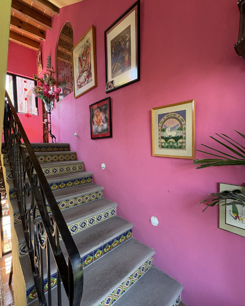 Interior of a house with pink walls decorated with framed artwork, a staircase with patterned tile risers, and a flower arrangement at the top of the stairs.