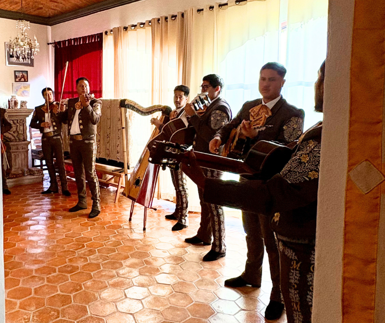 Five men dressed in traditional Mariachi outfits playing musical instruments including violins, guitars, and a harp in a room with hexagonal tile flooring and colorful curtains.