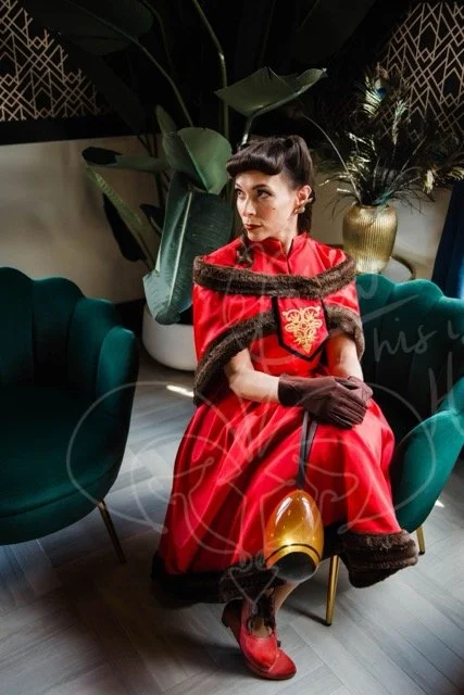 Hair in victory rolls with rolled bangs, she sits gazing to the side in a red dress with a mandarin collar and full skirt trimmed in brown fur. The off-the-shoulder capelet is also trimmed in brown fur with a gold swirl embroidered short tabard.