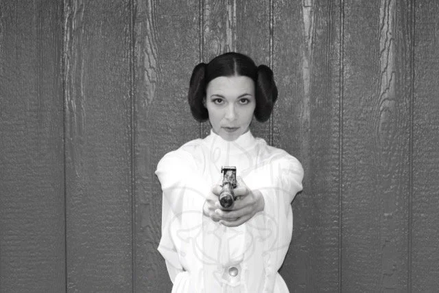 In black and white, Princess Leia aims her blaster at the camera while staring directly at you, iconic buns on either side of her head.