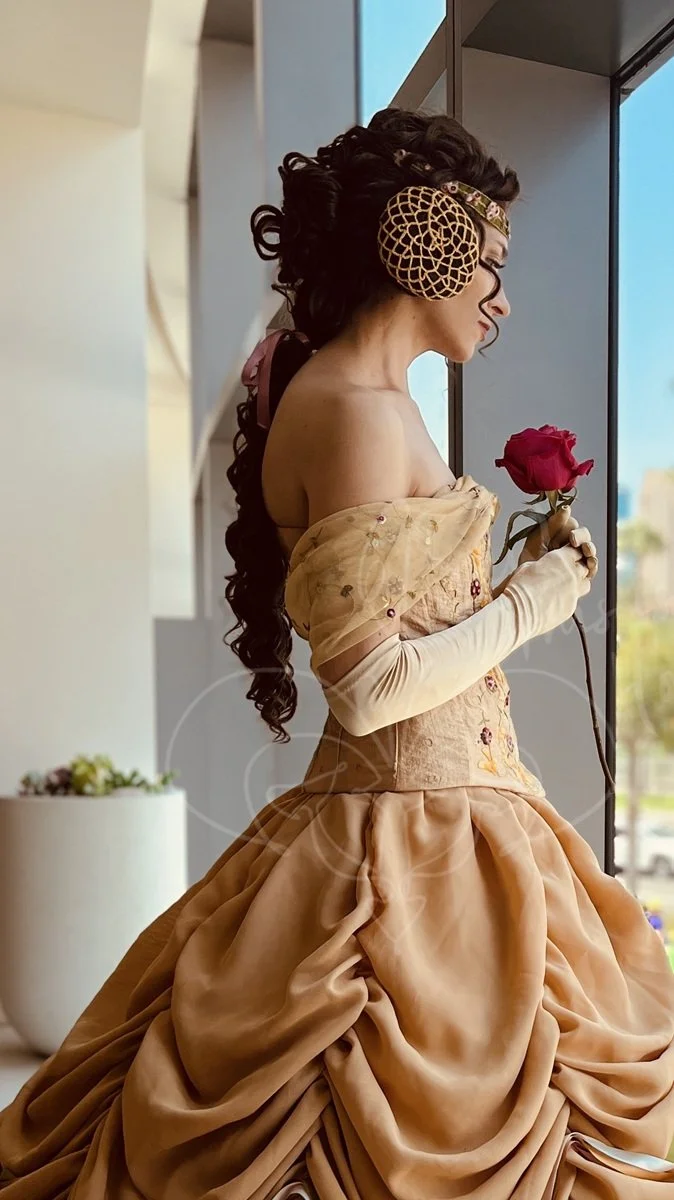 Dressed in a full dark gold skirt with an embroidered gold corset, a sheer embroidered off-the-shoulder gold shawl, and gold gloves, she looks down out a window holding a red rose.