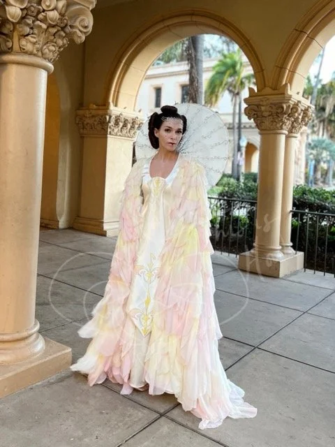 Standing under ornate columned arches, she is wearing an ivory satin gown with a tabard featuring a hand painted stylized Naboo symbol in pinks and yellows, which match the petals covering the full length cape. At her neck is a parasol type adornment