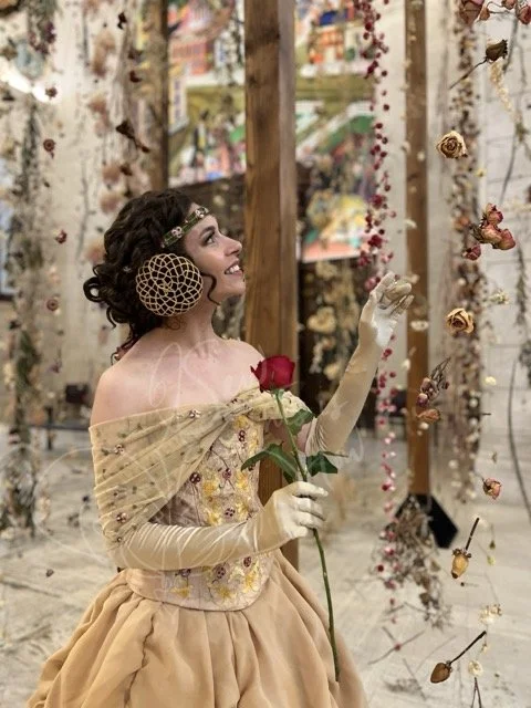 Wearing elbow length gold gloves and holding a rose in her right hand, she is reaching with her left hand toward strings of dried roses hung on invisible line as she looks up toward it. She wears a rose embroidered green headband and gold corset.
