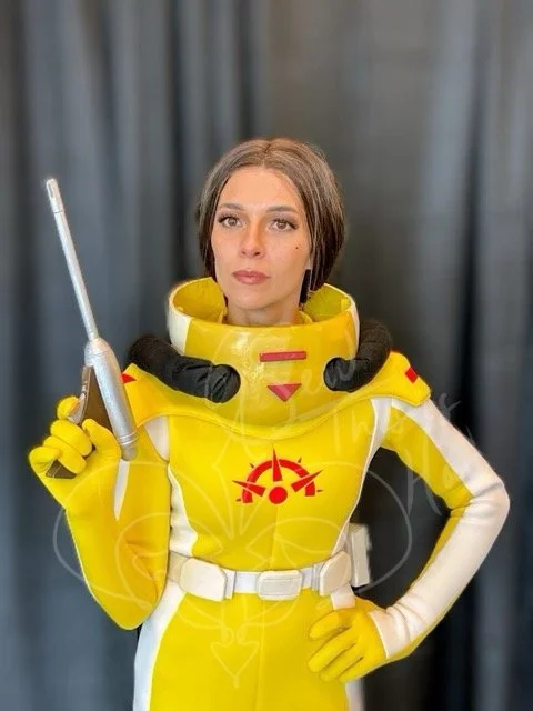 Shown from torso up in handmade yellow and white suit with red hazmat insignia on the breast. She is posed with left hand on hip, right hand holding a long barreled ELG-3A pistol.
