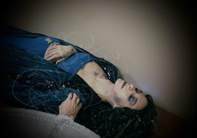 A hand reaches out to a "dead" Padmé, who lays in a blue gown and teal sequined cape with her hands across her belly holding the jappor snippet pendant. Her hands, neck, and face show dark markings of decay.