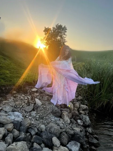 On a rocky shore with a bright sunset behind her, she looks down toward the water as she brings her arms to the side to catch the long flowing sleeves as the wind blows her airy medieval gown dyed in an ombre of cream, yellow, pink, and purple.