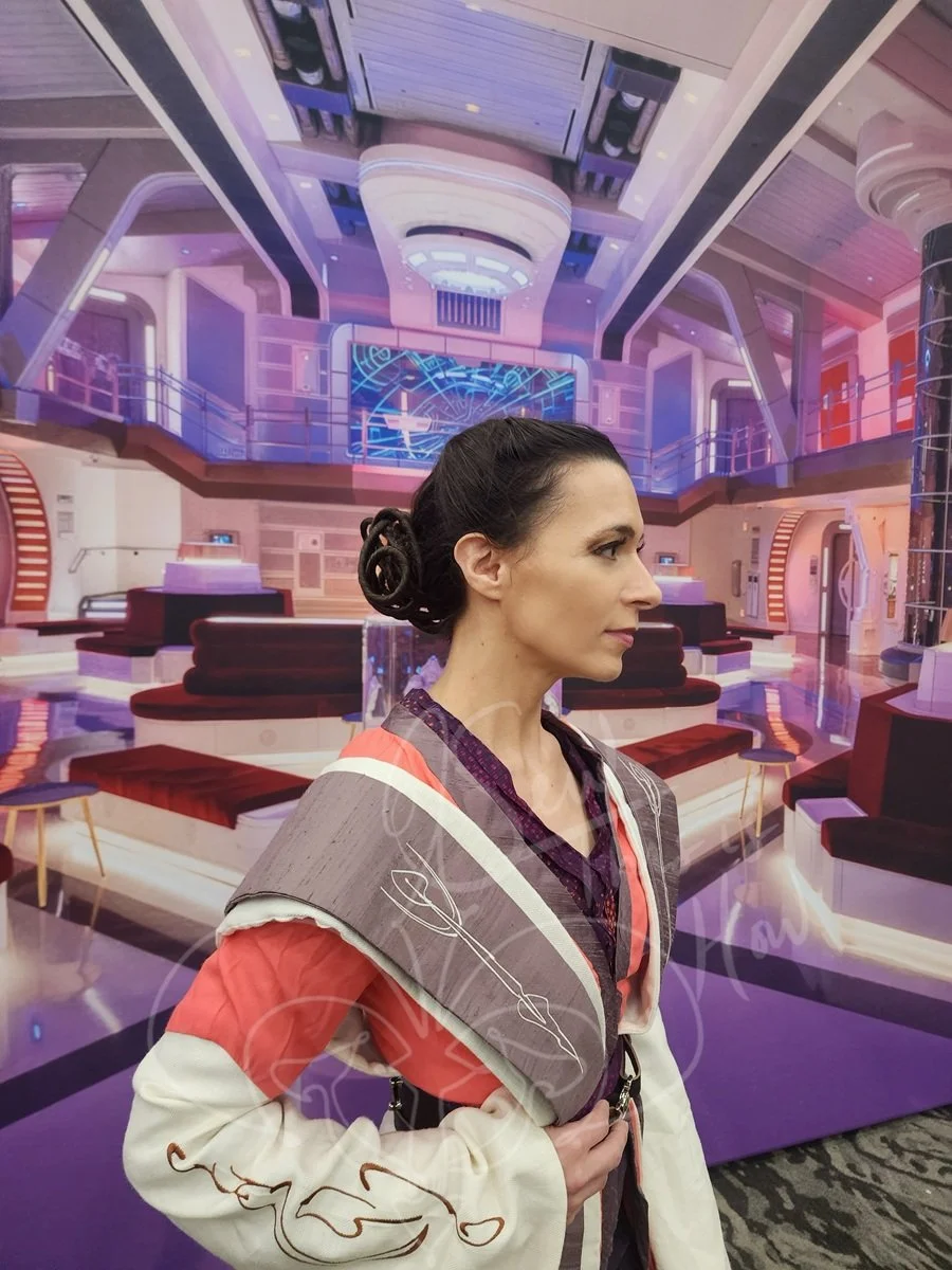 In front of a colorful purple-hued galactic transport background, she stands in dusty purple over ivory jedi tabards over a coral outer tunic and an aubergine inner tunic. The lower sleeves and outer tabards are embroidered with concept art motifs.