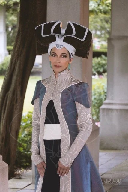 She wears a bone-colored headdress that encircles her brow and appears to support her brown hair up over a structure in the back, where it falls in a long fringe. The gown is deep navy with a grey-blue/red iridescent overcoat with swirling taupe trim