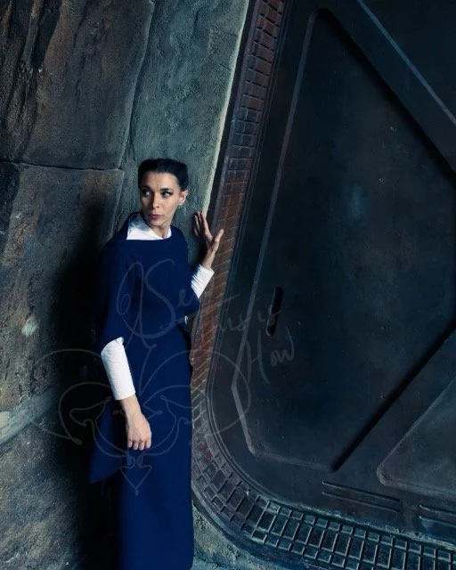 With one hand on the asymmetrical doorframe of a Star Wars set, she looks over the other shoulder wearing a tailored navy suit with a wide boatneck and a long skirt over a white long-sleeved mock turtleneck.