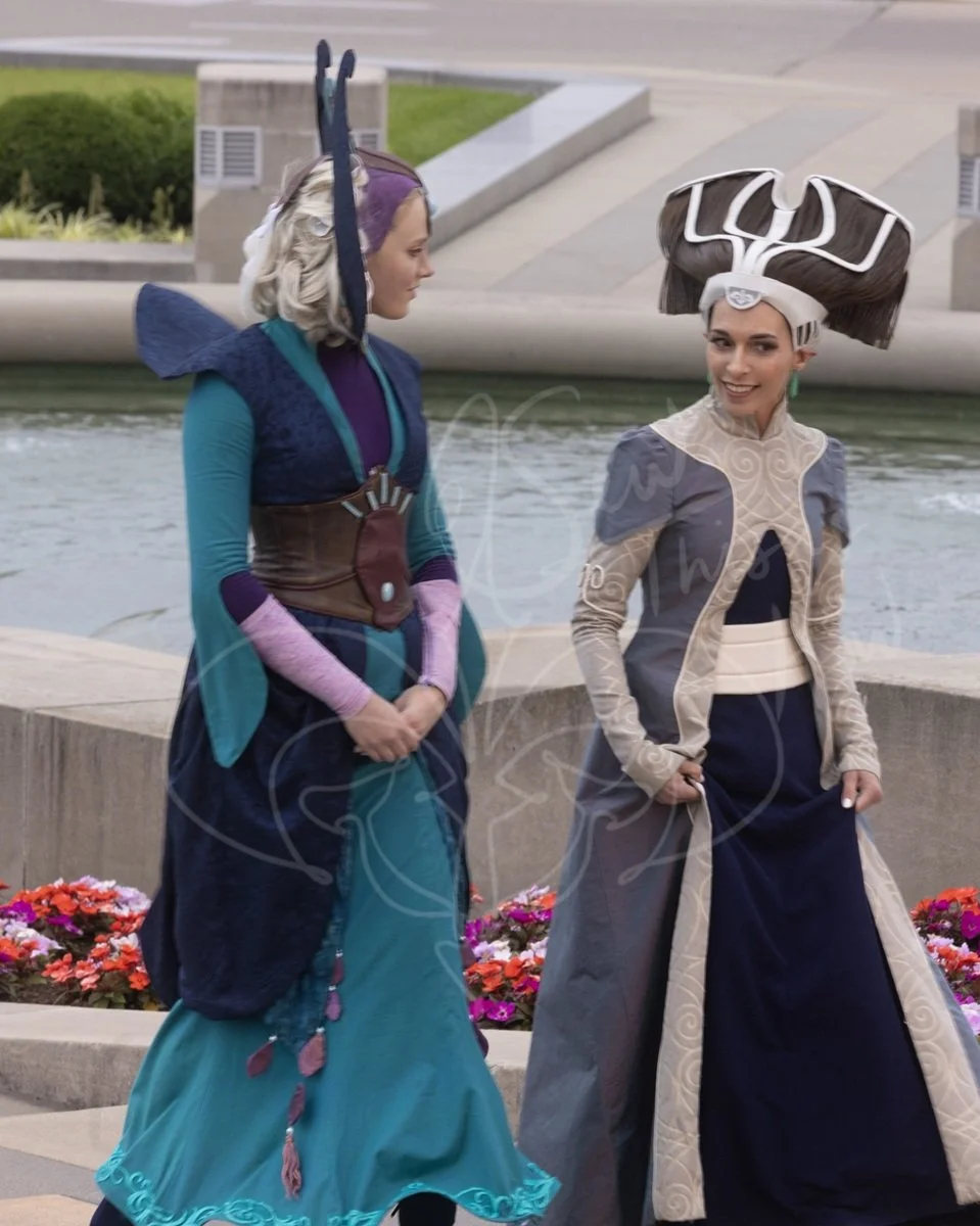 Walking in front of an unban water fountain lined with flowers, she wears her animated Clone Wars senate gown while chatting with a colleague dressed as Duchess Satine of Mandalor.